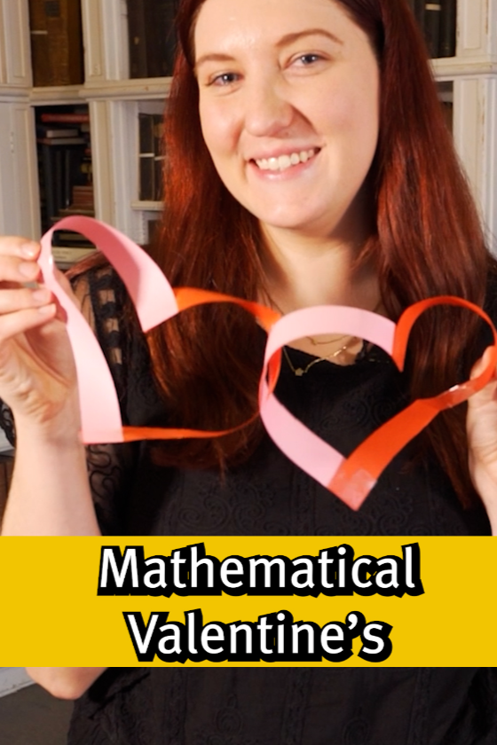 A woman with long red hair smiling and holding a paper heart-shaped craft made of pink and red paper, with a yellow banner at the bottom that reads "Mathematical Valentine's" in bold black and white text.