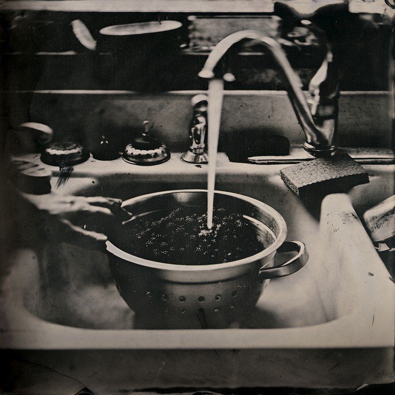  A black and white photo of a sink and faucet with a colander full of berries. 