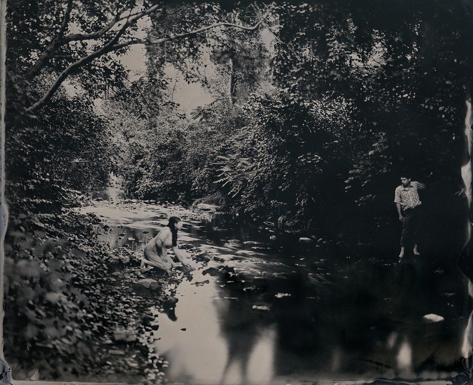  A black and white photo of a couple on a river in a forest. 