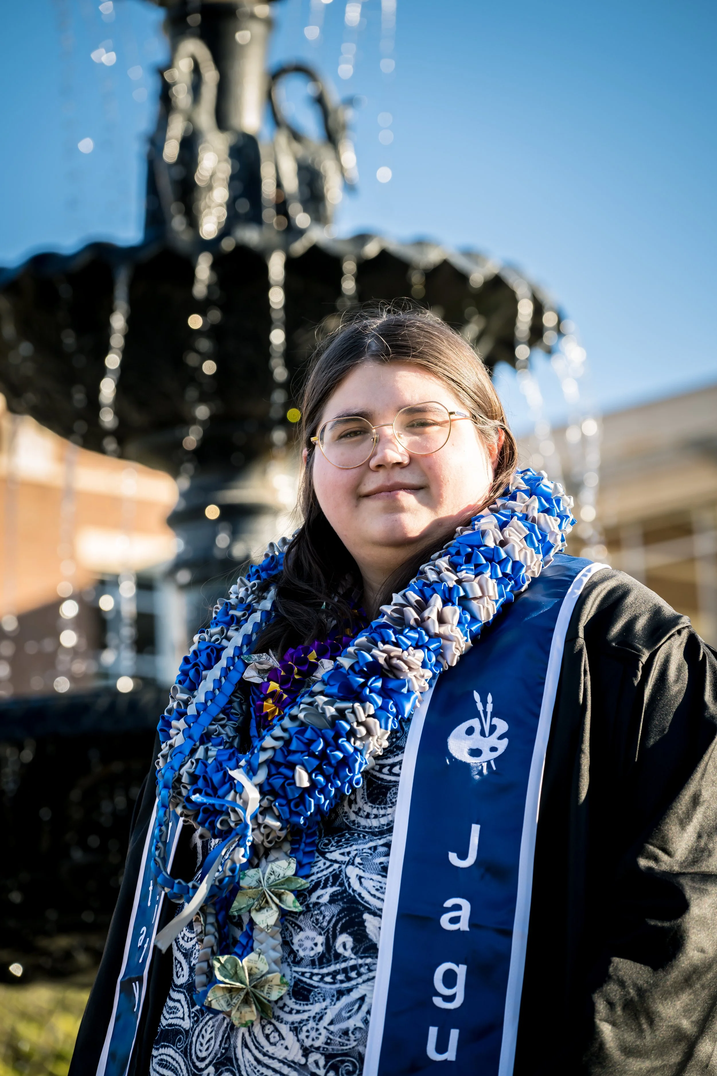 A young woman with glasses and long dark hair standing outdoors in front of a decorative fountain. She is wearing a blue sash with white lettering, a patterned dress, and a blue and gray ribbon lei around her neck. She is smiling softly at the camera