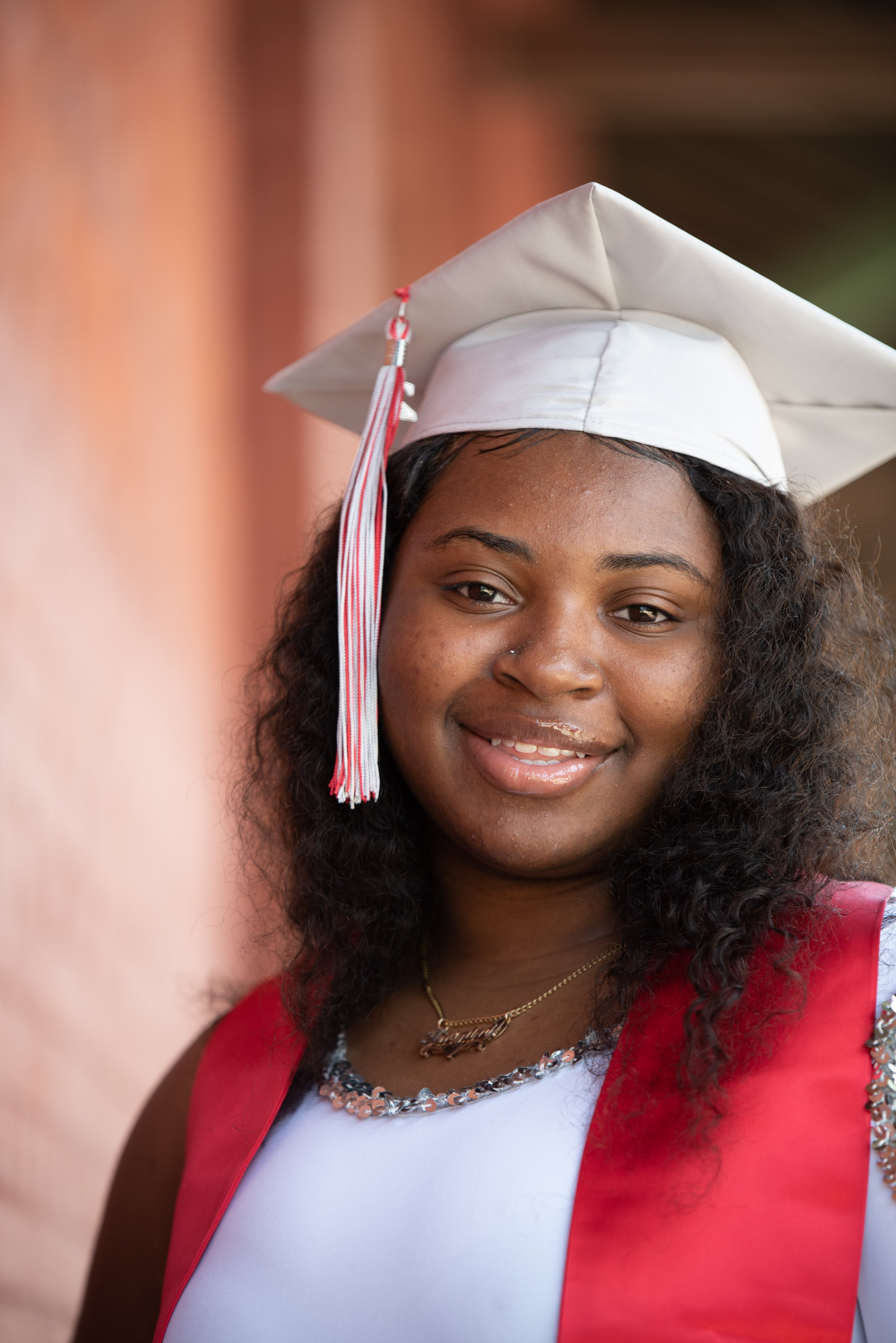 A young woman with curly hair wearing a white graduation cap with red and white tassel, a white shirt, a red vest, and a gold necklace, smiling.
