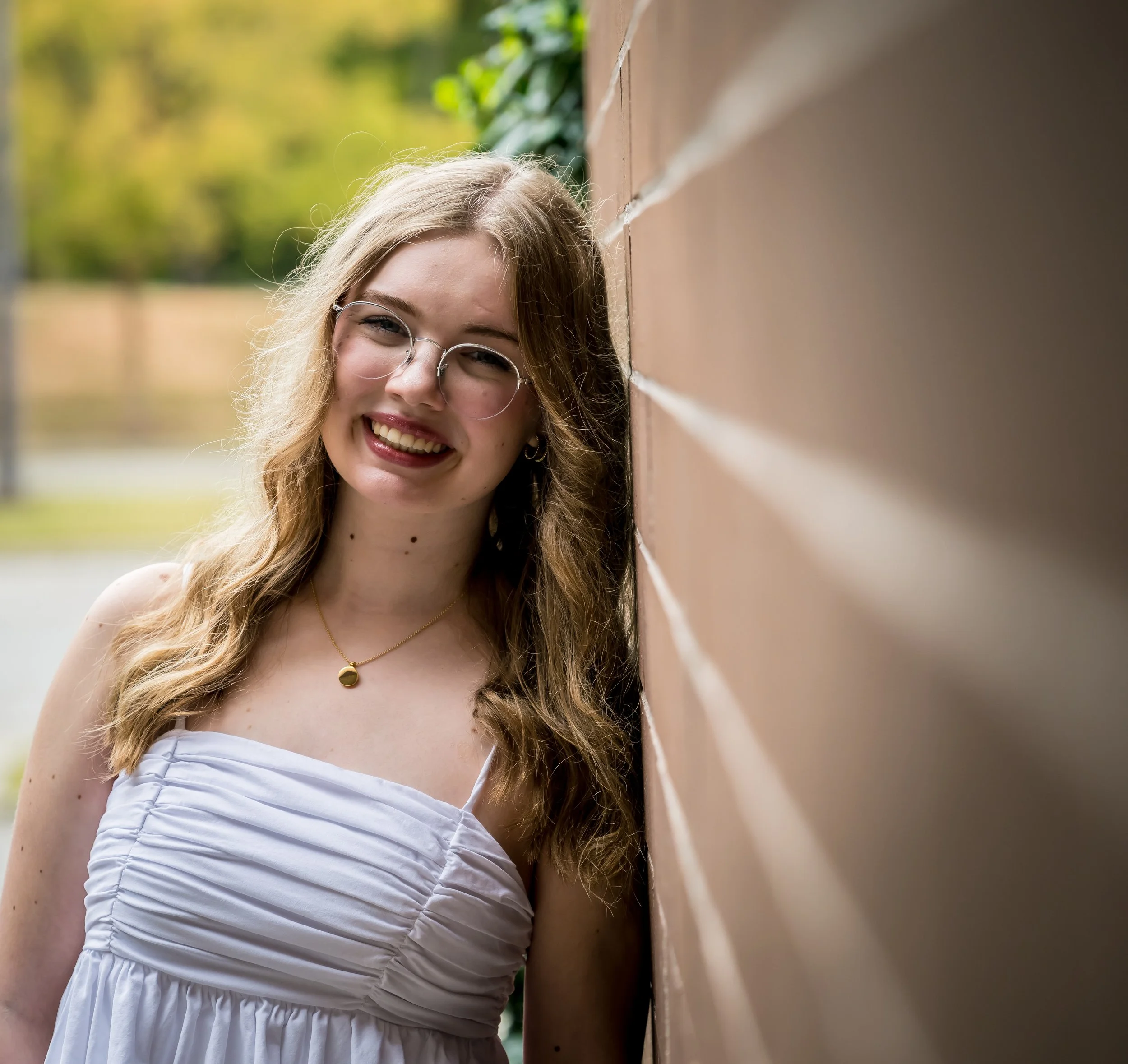 A young woman with long, wavy blonde hair and glasses smiling while leaning against a brick wall outdoors, wearing a white sleeveless dress with ruched details.