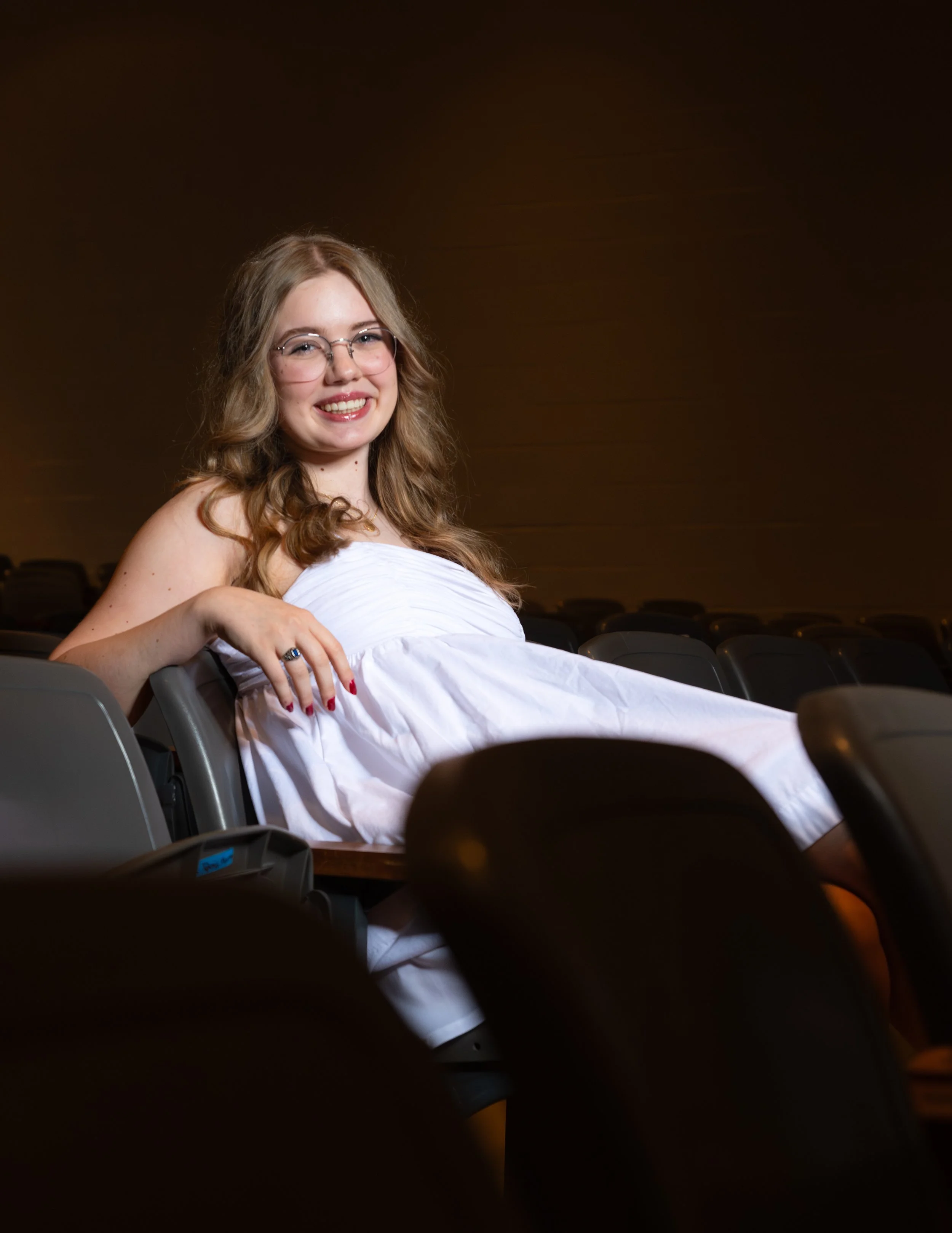 Young woman with glasses smiling and lying on her side on theater seats, wearing a white dress.