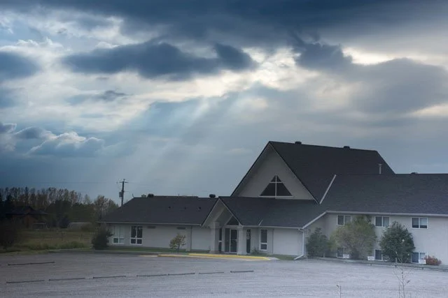 The Okotoks Alliance Church exterior and parking lot under a cloudy sky with sun rays in the background.