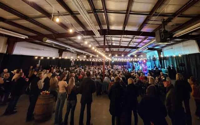 A live music event with a large audience in front of a colourfully-lit stage in the Legacy Hall at the Cochrane Lions Club Events Centre.