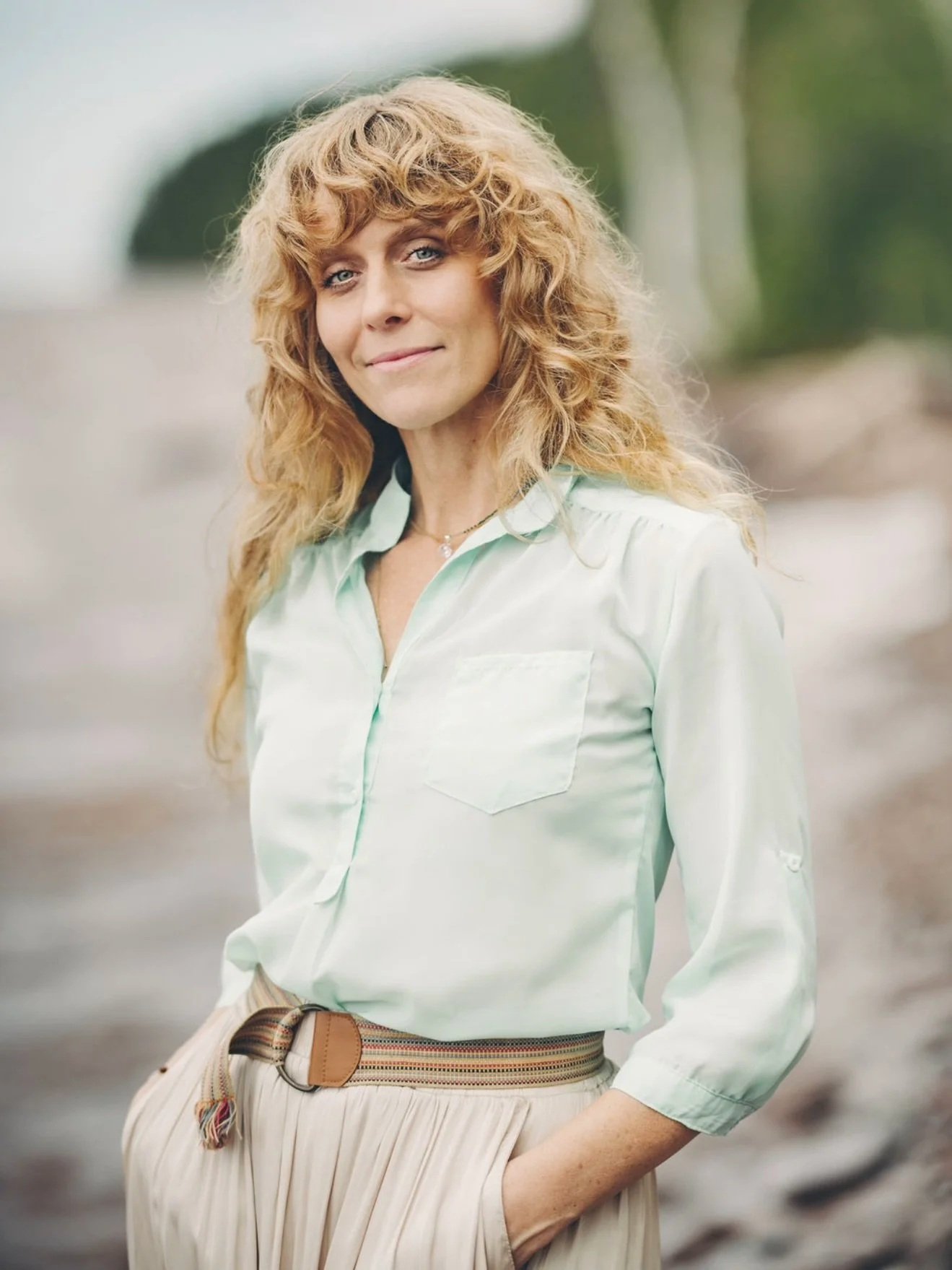 A woman with curly blonde hair wearing a light green button-up shirt and beige pleated pants, standing outdoors with a blurred background.