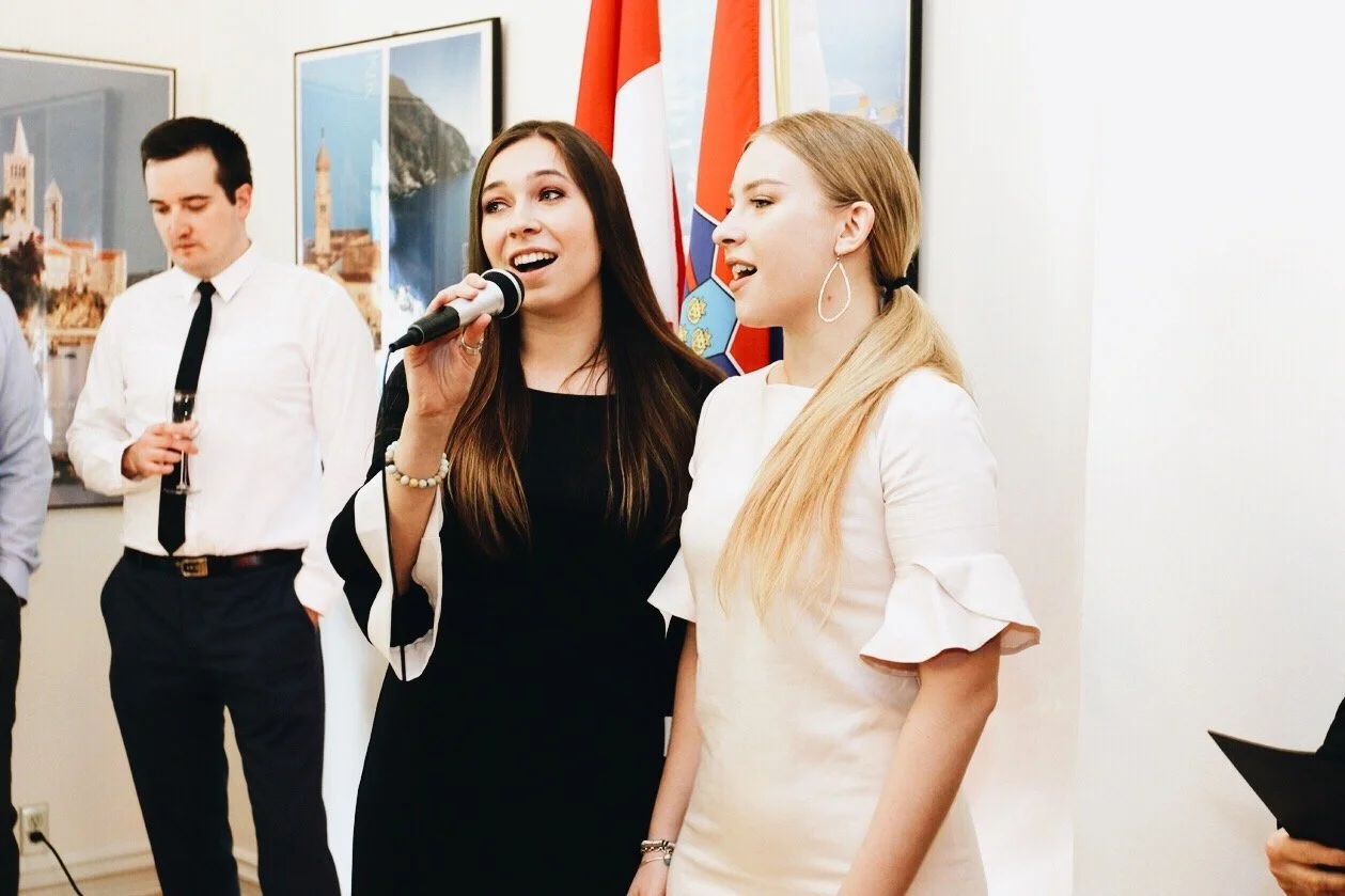 Two women speaking into a microphone at a formal event, with flags and framed pictures in the background, and a man holding a drink nearby.