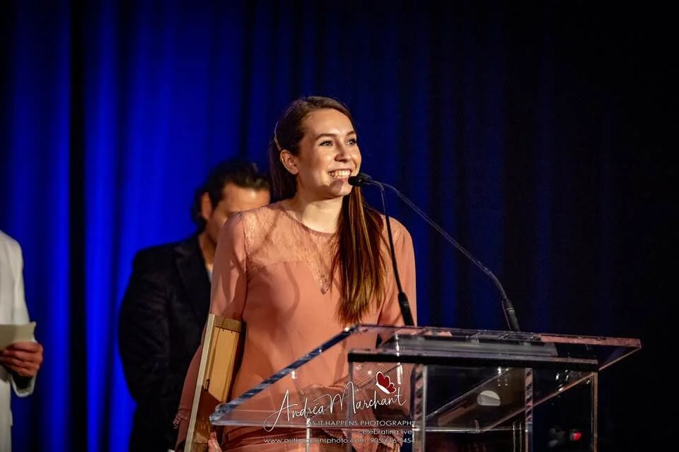 A woman with long brown hair smiling while speaking at a podium during an event, with blue curtains in the background.