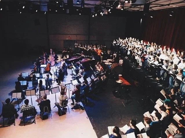 An orchestra and choir rehearsing in a dark concert hall with music stands, chairs, and musical instruments.