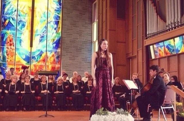 A young woman singing on stage with a choir and a musician playing guitar beside her at a concert hall with colorful stained glass windows.
