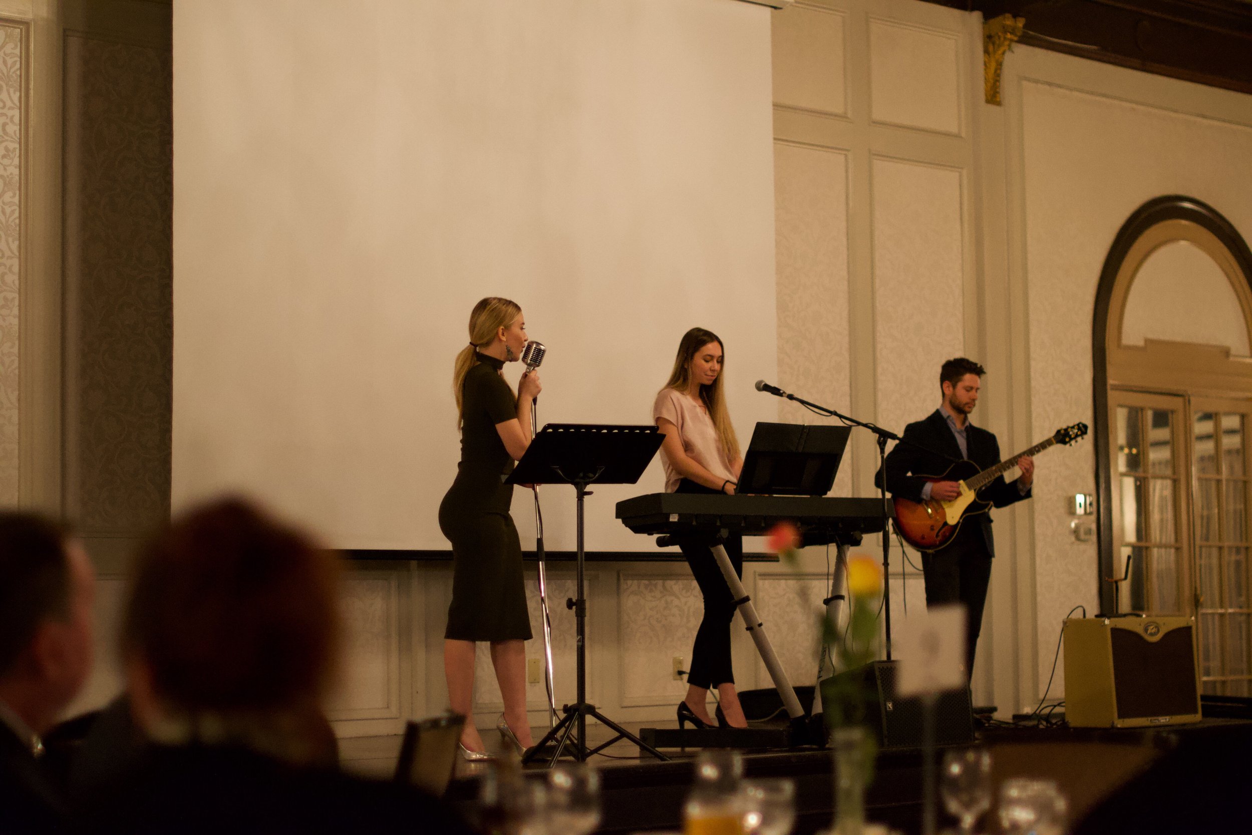 Three musicians performing on a stage: a woman singing into a vintage microphone, a woman playing keyboard, and a man playing guitar, in a formal setting. Dani Kristina, Andrew Sporcic, Julijana Hajdinjak. 
