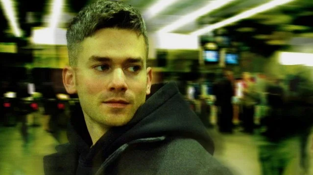 Young man with short brown hair and light skin, wearing a black hoodie, standing in a busy indoor public space. Singer Matt Ryan.
