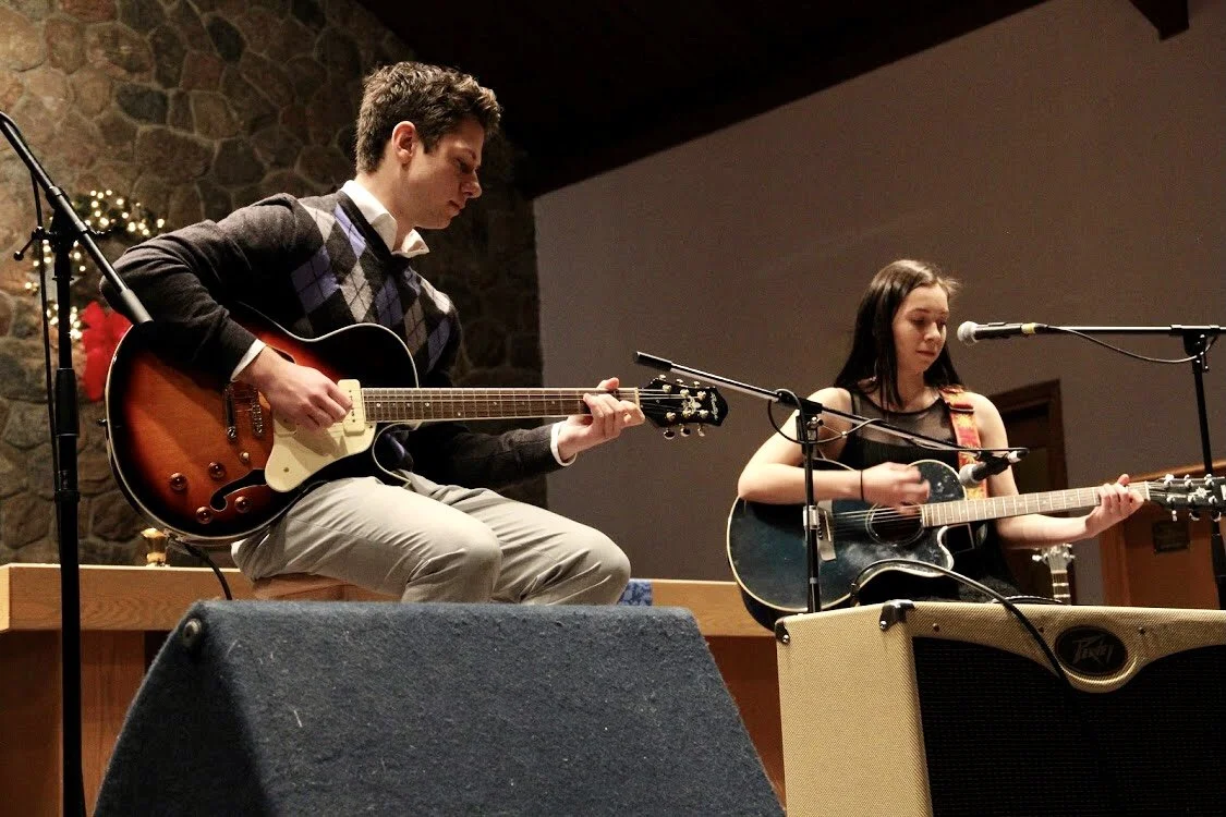 A young man and young woman playing acoustic guitars on stage with microphones in front of them, during a performance, with a stone wall and a decorated background.