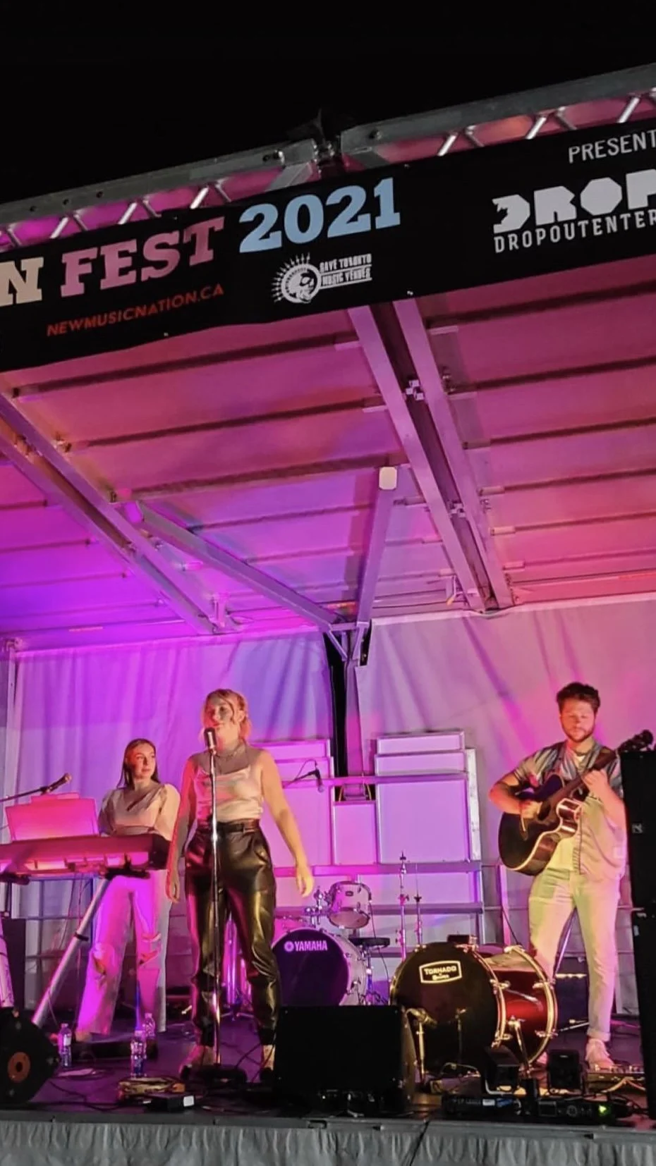 Band performing on stage at outdoor music festival with pink lighting, featuring female singer with a microphone, keyboardist, drummer, and guitarist, under a banner for the 2021 Music Fest.