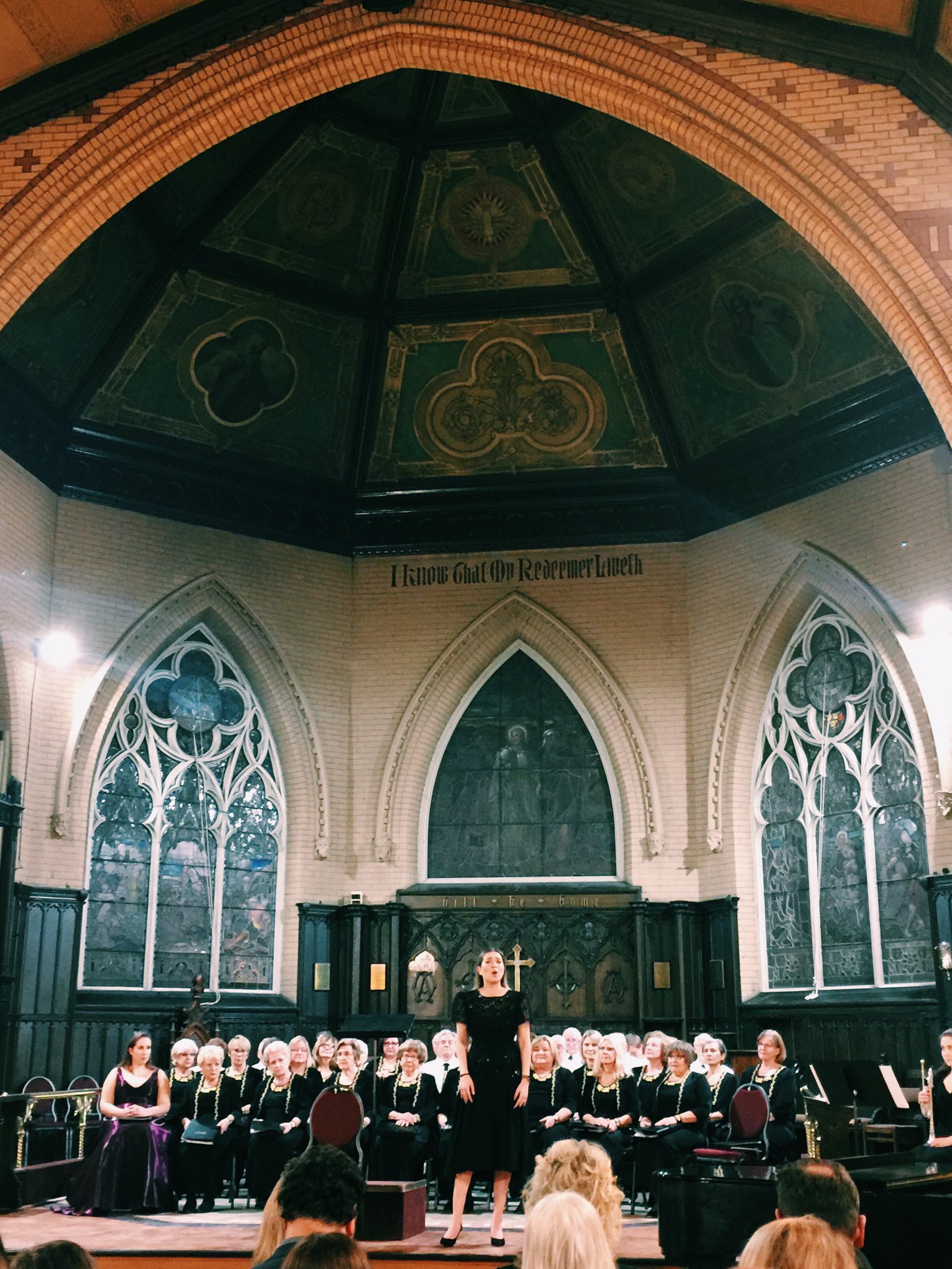 A choir performing on stage inside a church with stained glass windows and a large dark wooden altar behind them. A woman in a black dress is singing in front of the choir, with audience members visible in the foreground.