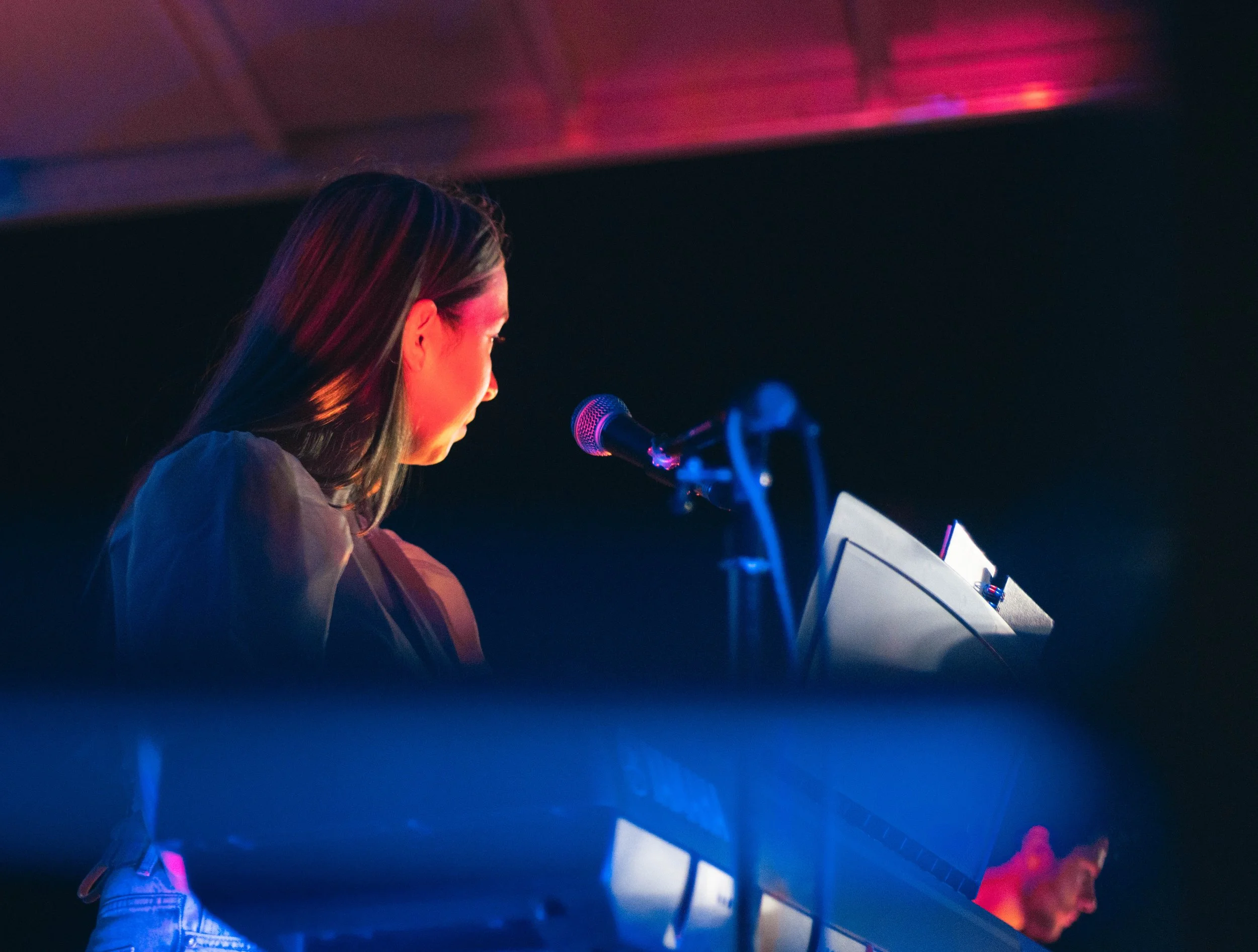 A woman with long dark hair singing or speaking into a microphone on stage, illuminated by colorful stage lighting, with a music stand and sheet music in front of her.