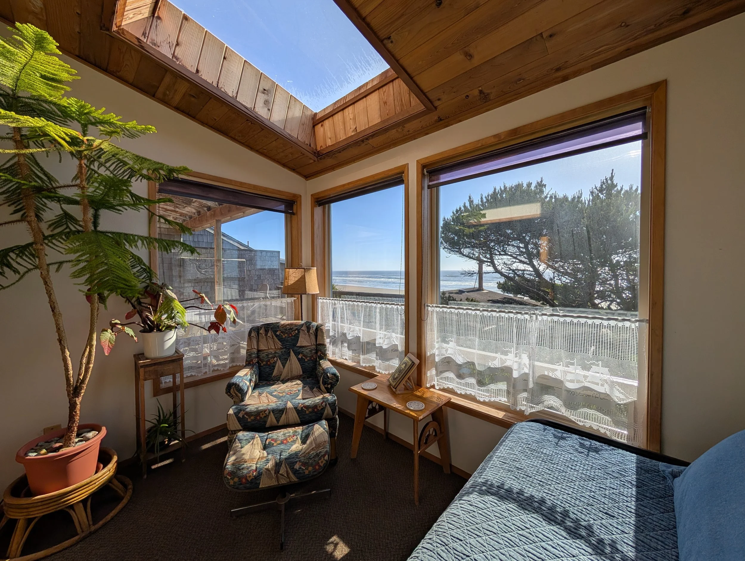 A sunlit living room with large windows showing a view of the ocean and trees. A patterned armchair with a matching ottoman is near the window, surrounded by potted plants. The ceiling features a skylight with wooden trim, allowing additional sunligh