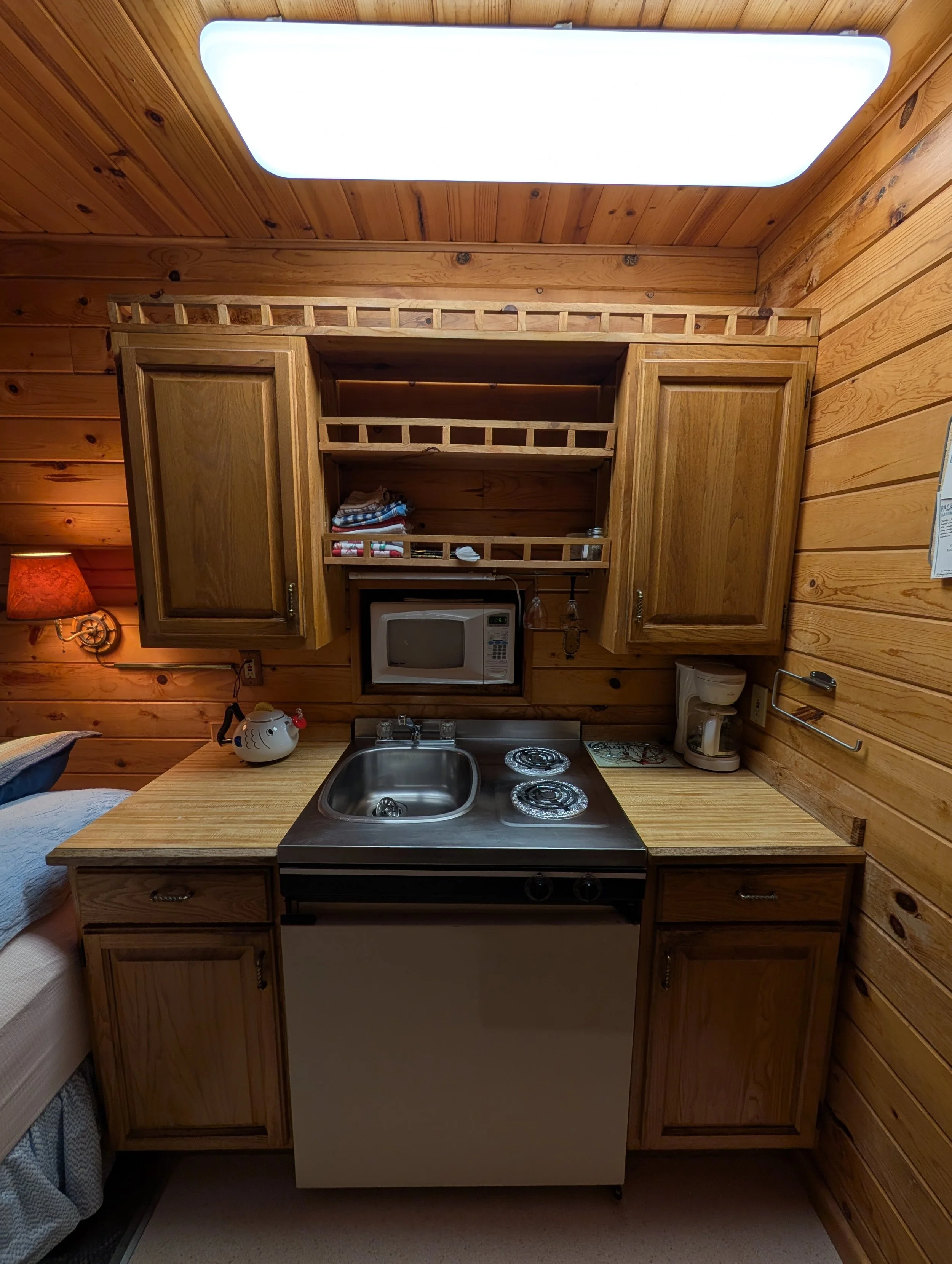 Wooden kitchen area with cabinets, a microwave, a small sink, a stovetop, and a countertop. The walls and ceiling are paneled with wood, and there is a bright ceiling light.