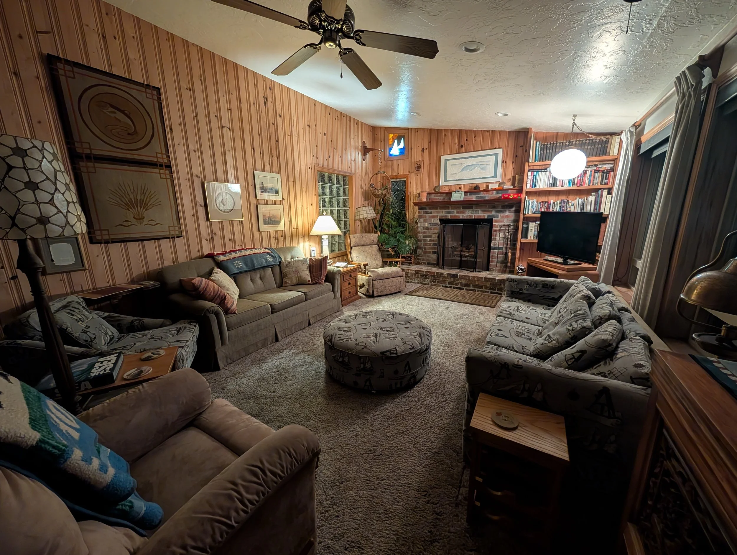 A cozy living room with wood-paneled walls, a brick fireplace, a flat-screen TV, and various sofas and armchairs with patterned and solid cushions. There are lamps, framed artwork on the walls, a bookshelf, and ceiling fans.