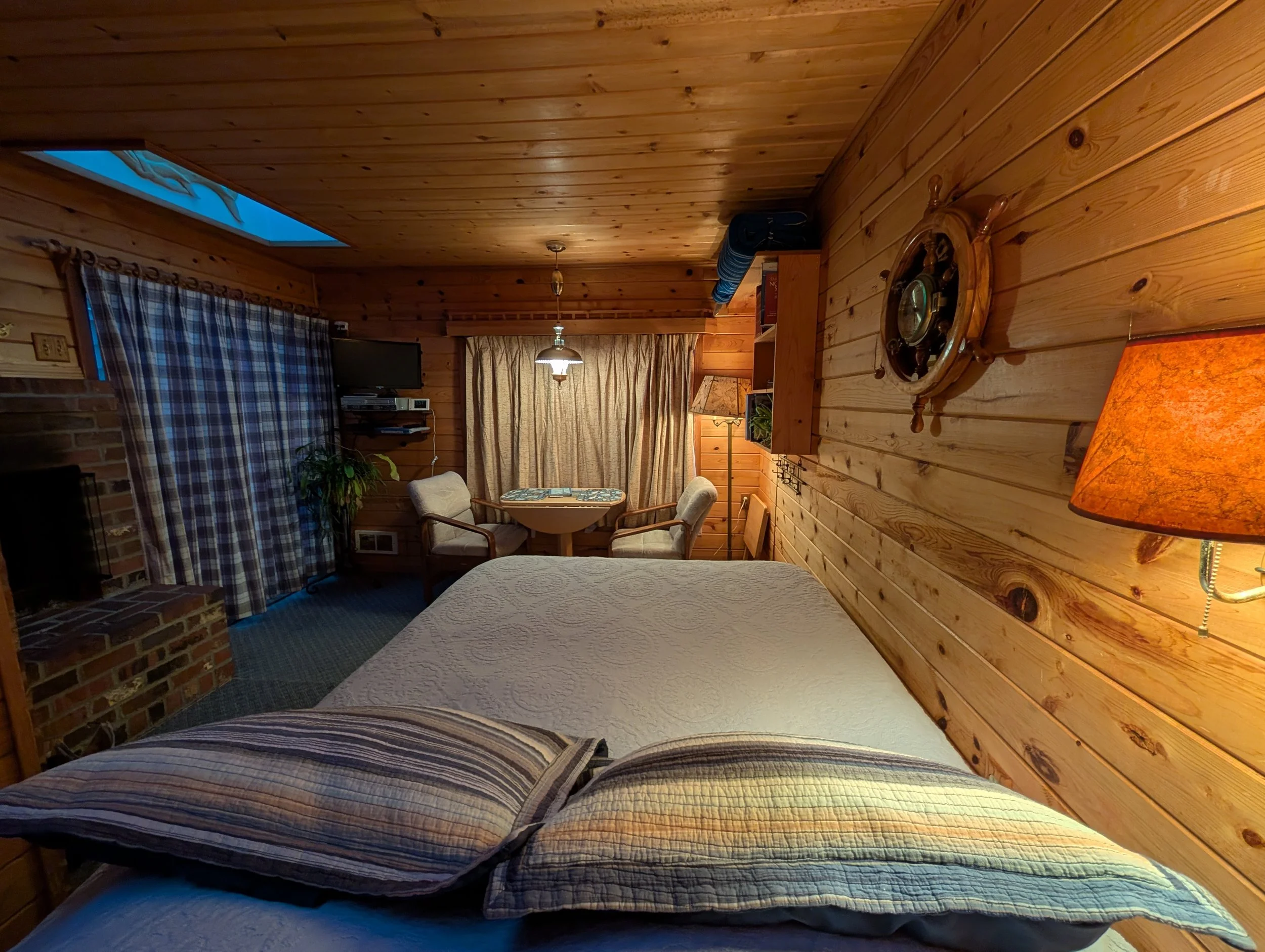 Interior view of a cozy log cabin room with wooden paneling, a dining area with a round table and chairs, a window with plaid curtains, and decorative nautical items on the wall.