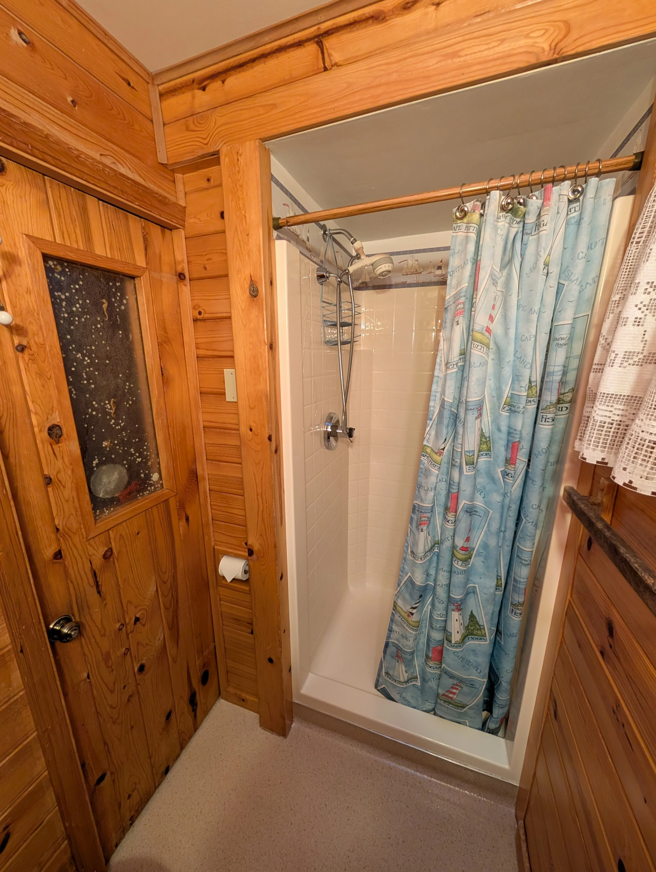 A bathroom with wooden walls and ceiling, beige tile shower with a colorful lighthouse-themed shower curtain. There is a showerhead with a hose and a metal rack inside the shower. Outside the shower, there is a window with a textured glass panel and 