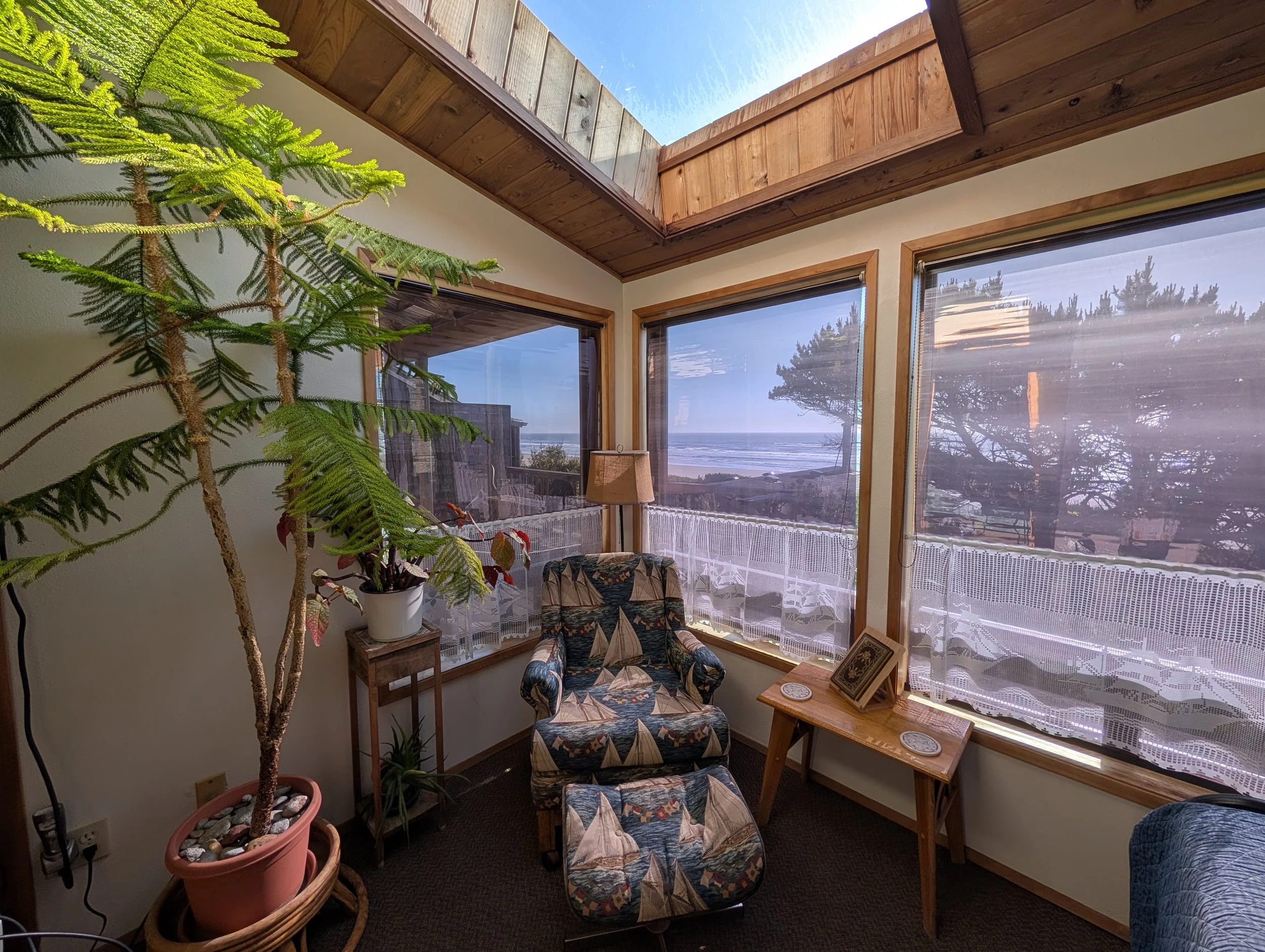 A cozy room with large windows overlooking the ocean, featuring indoor plants, a floral-patterned armchair, side tables with coasters, and a wooden table with picture frames, all decorated in warm, natural tones.