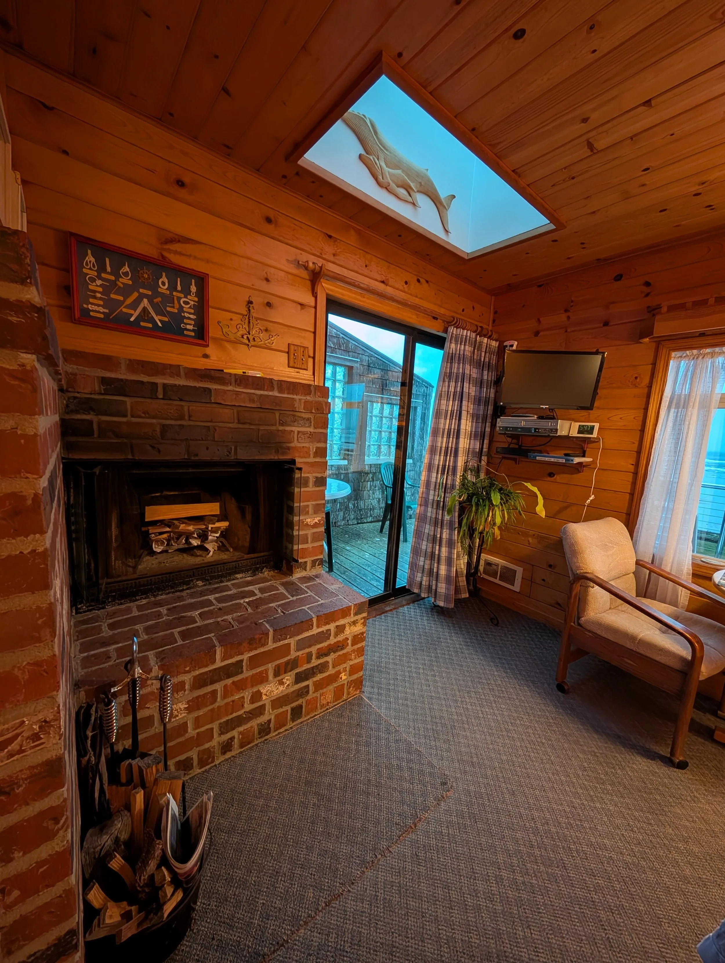Interior of a cozy room with wooden walls and ceiling, a brick fireplace, a sliding glass door with plaid curtains leading to an outdoor area, a television mounted on the wall, a chair, and a potted plant. There is a skylight with a decorative dolphi
