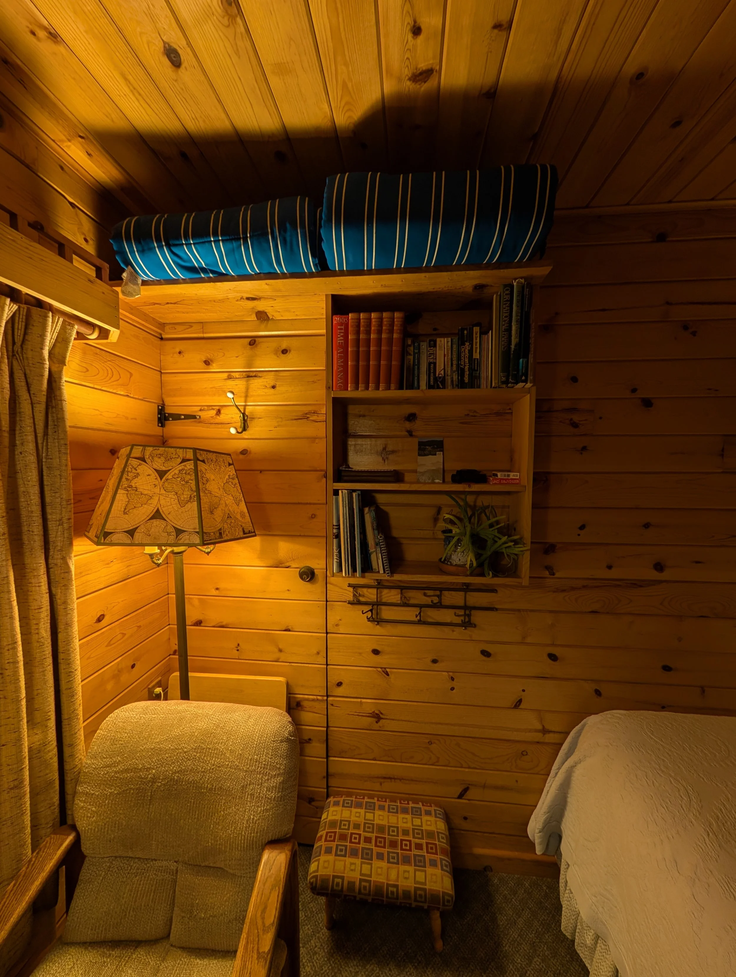 Interior of a cozy wooden room with a wooden bookshelf, a lamp with a world map lampshade, a chair, a small patterned footstool, a bed with a light-colored blanket, a wall-mounted coat rack, and a window with a curtain.