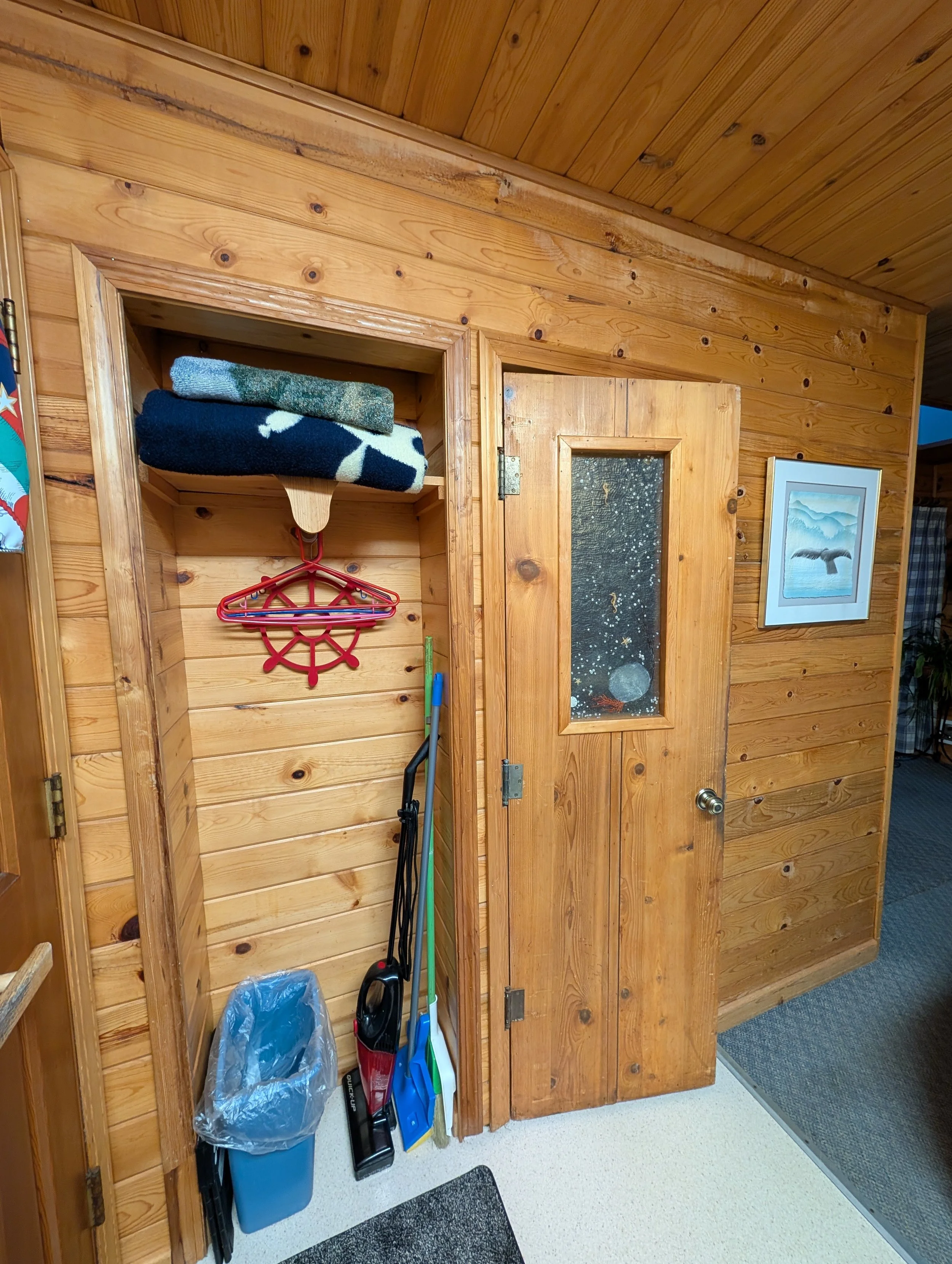 A wooden door and wall in a room with some cleaning supplies and a garbage can.