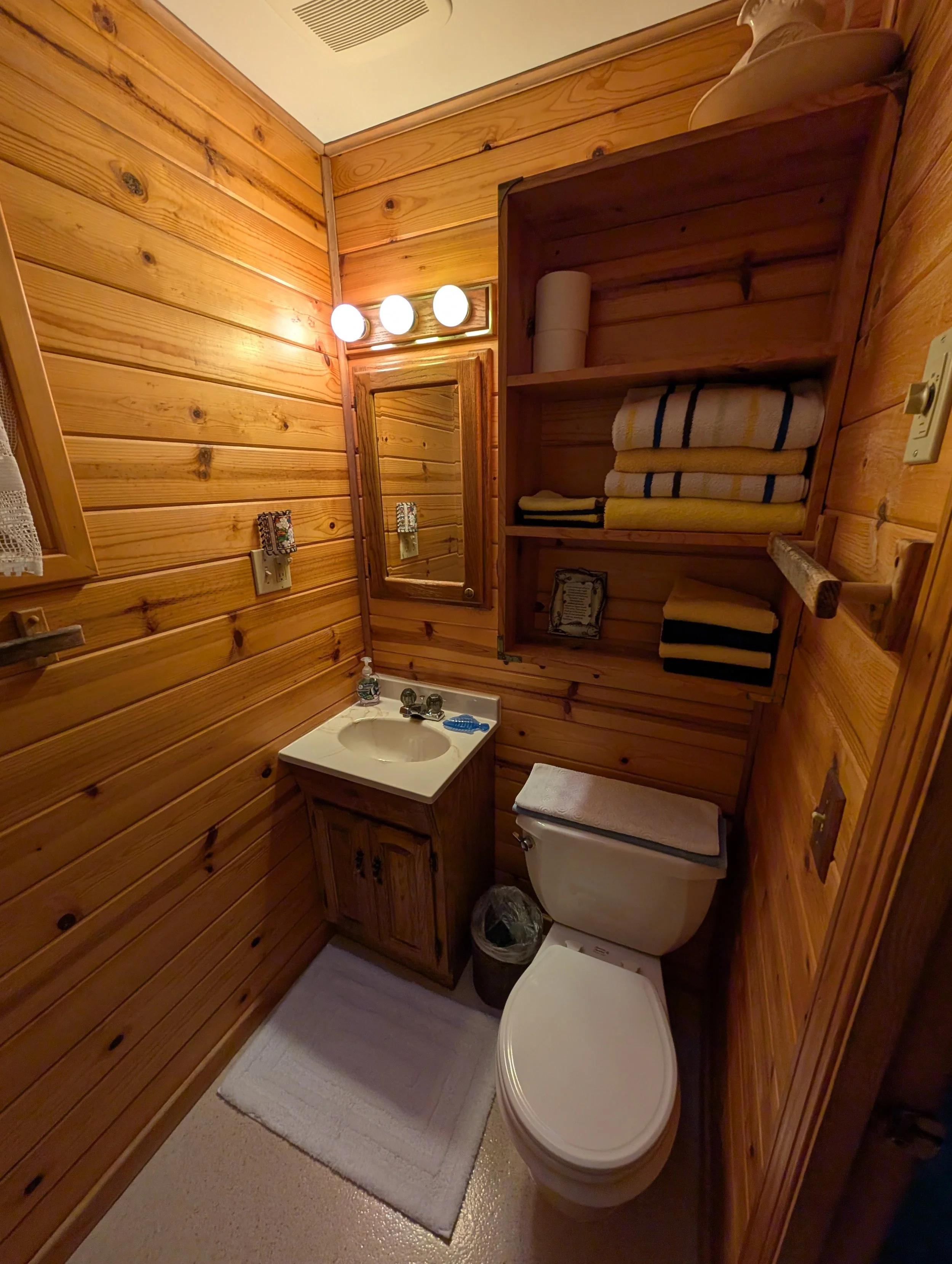 A small rustic bathroom with wood-paneled walls, a mirror above a small sink, and a toilet with a towel on top. There is a cabinet with folded towels and shelving. A light fixture with three bulbs is above the mirror, and there is a small window with