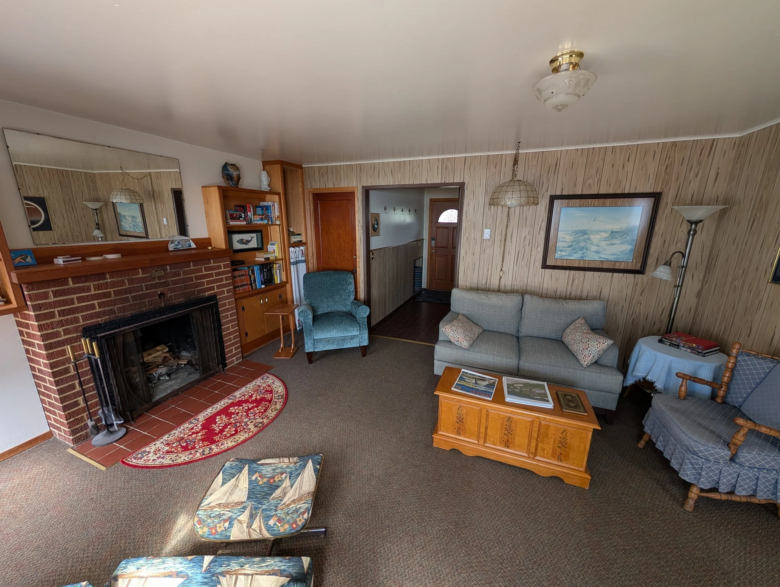 Living room with brick fireplace, wooden bookcase, blue patterned armchairs, beige sofa, coffee table with magazines, wall art, and wooden door.