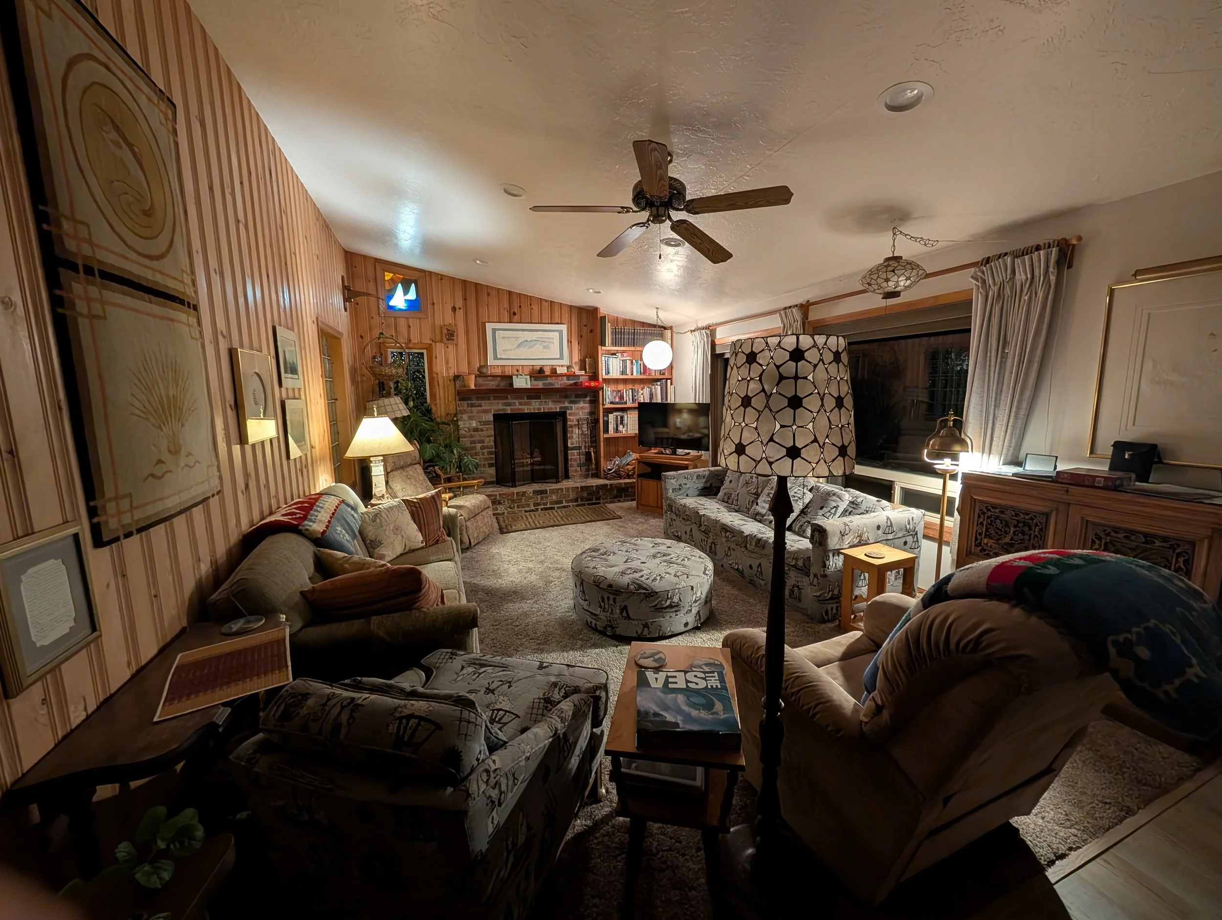 Living room with wood-paneled wall, brick fireplace, bookshelf, ceiling fan, and various couches and chairs with patterned upholstery.
