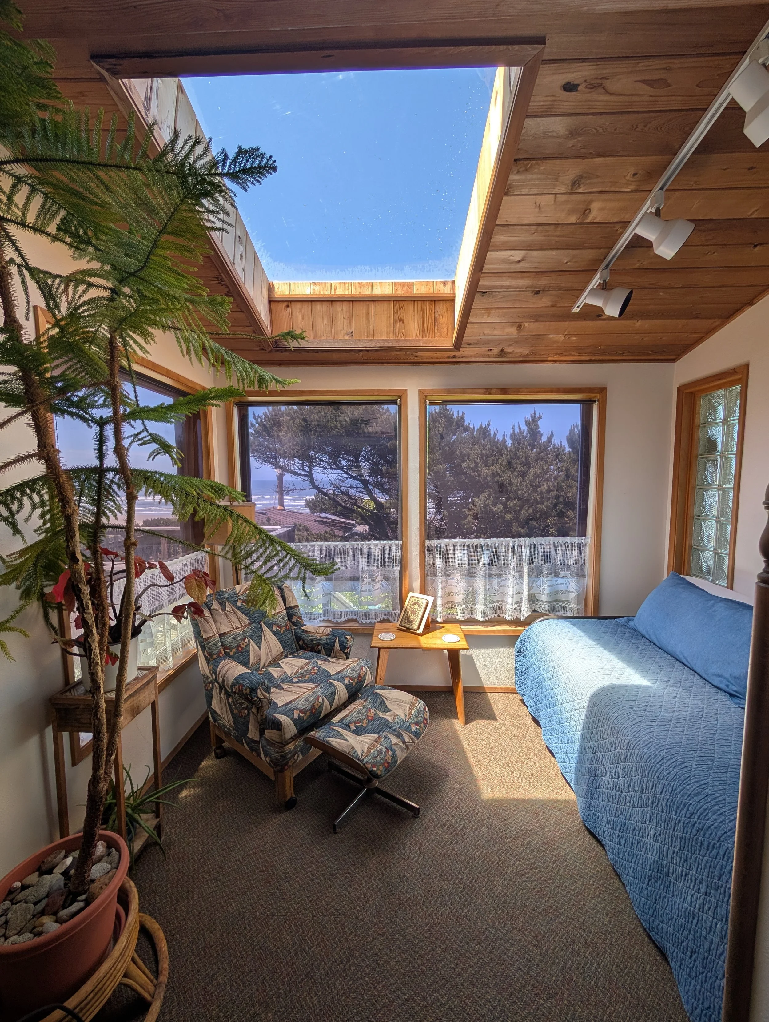 Living room with large windows, a blue quilted couch, a floral-patterned armchair, a small wooden side table with picture frames, potted plant, and a skylight in the wooden ceiling.