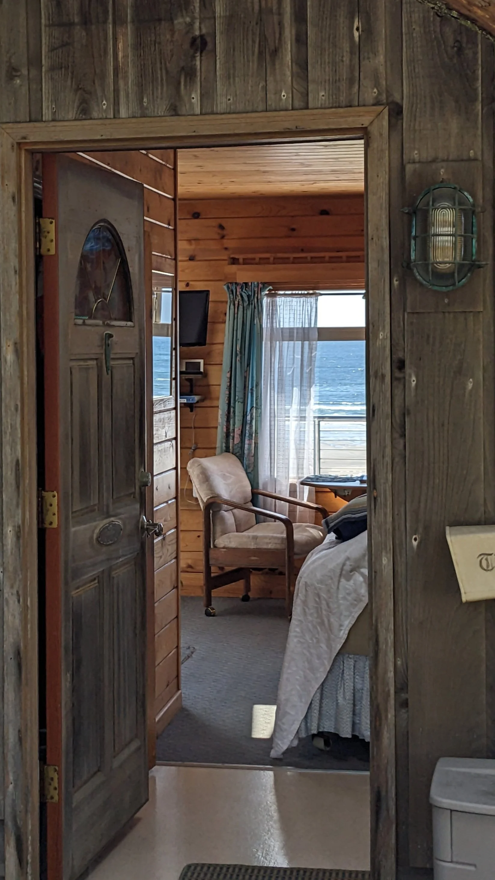 View through a rustic wooden doorway into a cozy bedroom with a view of the ocean outside. The room features wooden walls and ceiling, a window with curtains, a chair, a bed, and a small TV on a shelf.