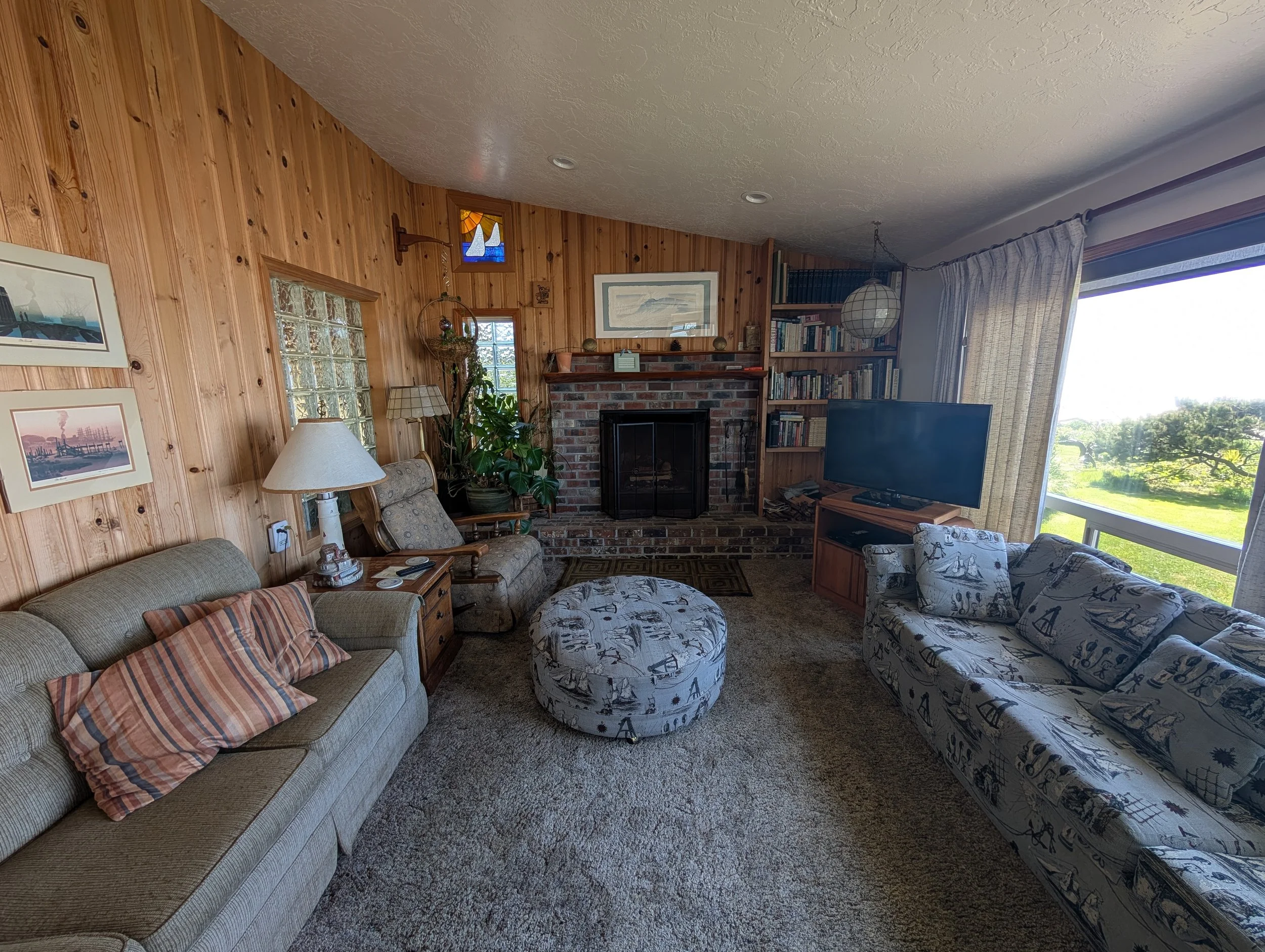 Living room with wood-paneled walls, a brick fireplace, a flat-screen TV, patterned couches, an ottoman, framed pictures, a lamp, a bookshelf, curtains, and large windows showing a green outdoor landscape.