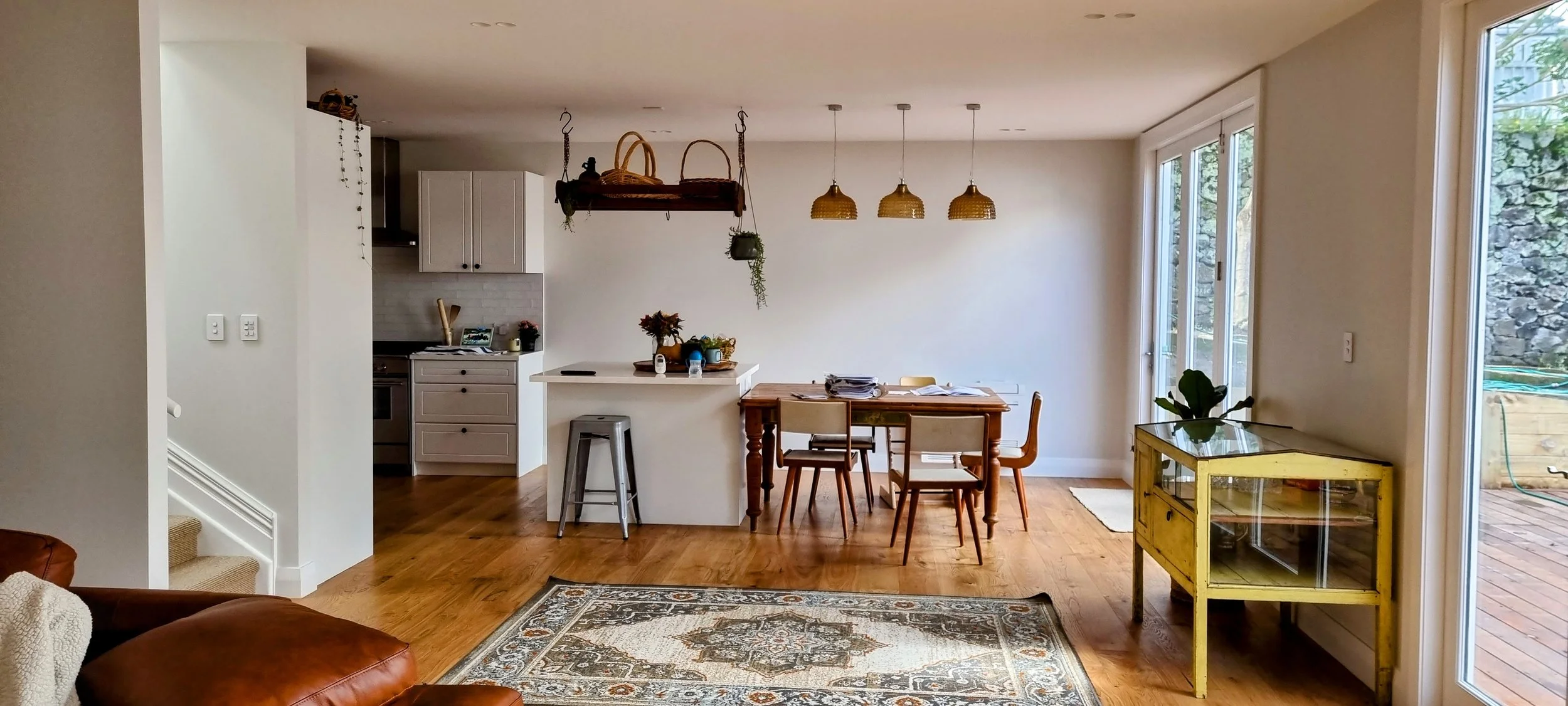 Living room and dining area with hardwood flooring, a brown leather sofa, a vintage yellow cabinet, a dining table with four chairs, hanging woven pendant lights, sliding glass doors to an outdoor deck, and a kitchen with white cabinets in the background.