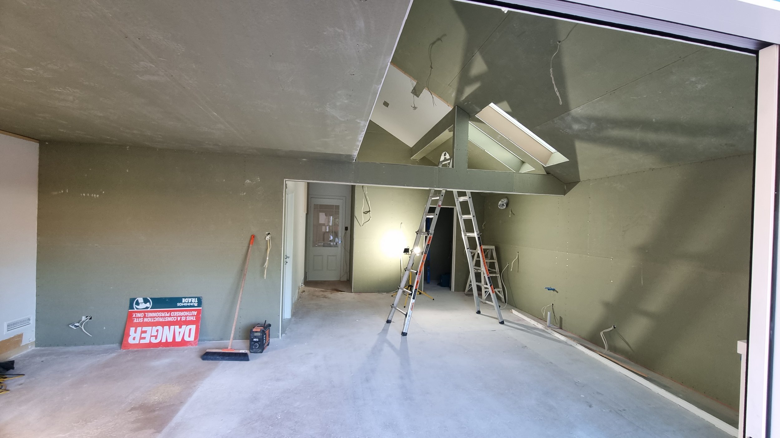 Interior of a room under construction with unfinished walls, ceiling, and floor. A ladder is positioned in the center, and construction tools and equipment are scattered around. There is an angled ceiling with a skylight and some exposed wiring.