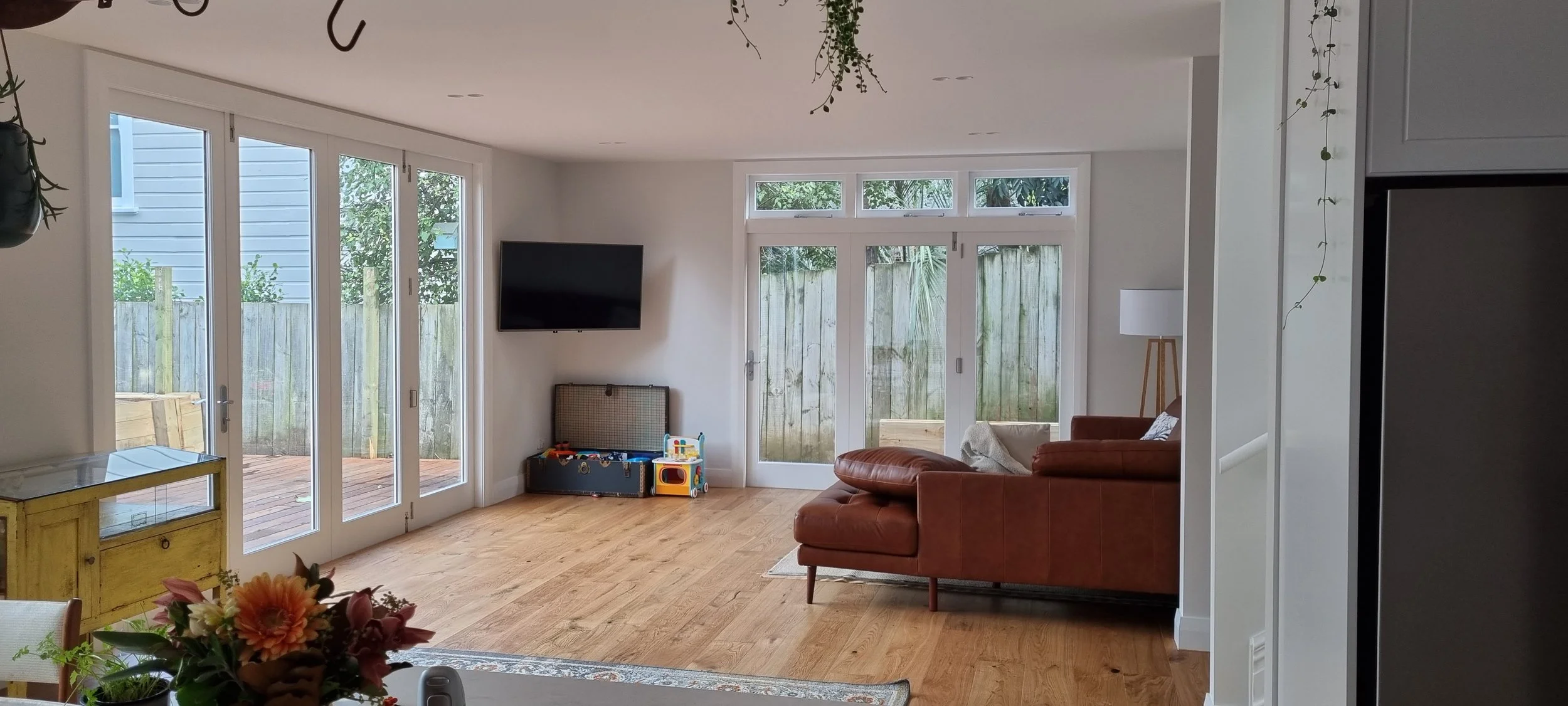 Living room with large glass sliding doors leading to backyard, brown leather sofa, wall-mounted TV, children's toy box, and floor lamp, with a wooden floor and outdoor fenced yard.
