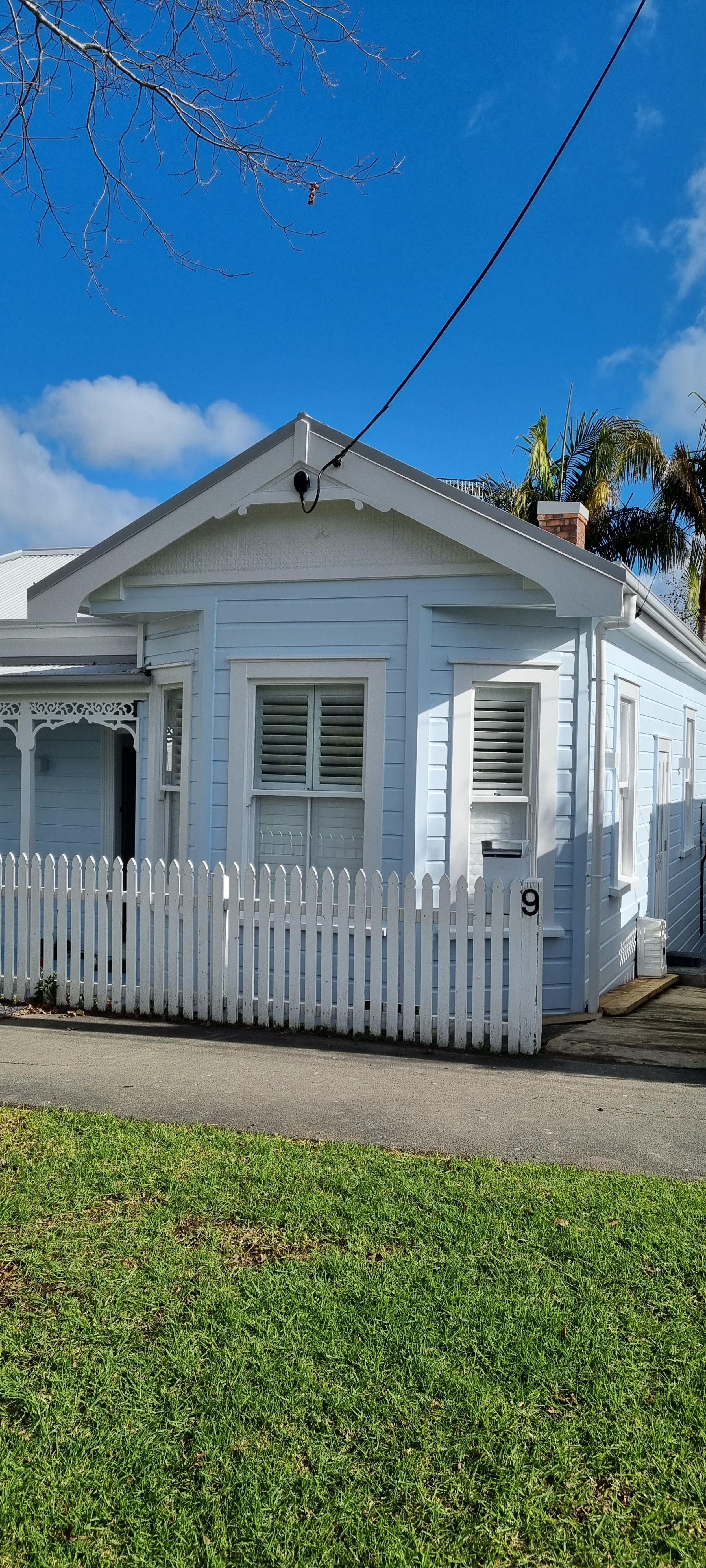 A white house with a picket fence, two windows, and a small porch, under a blue sky with a few clouds. A tree with bare branches is visible in the corner of the image.