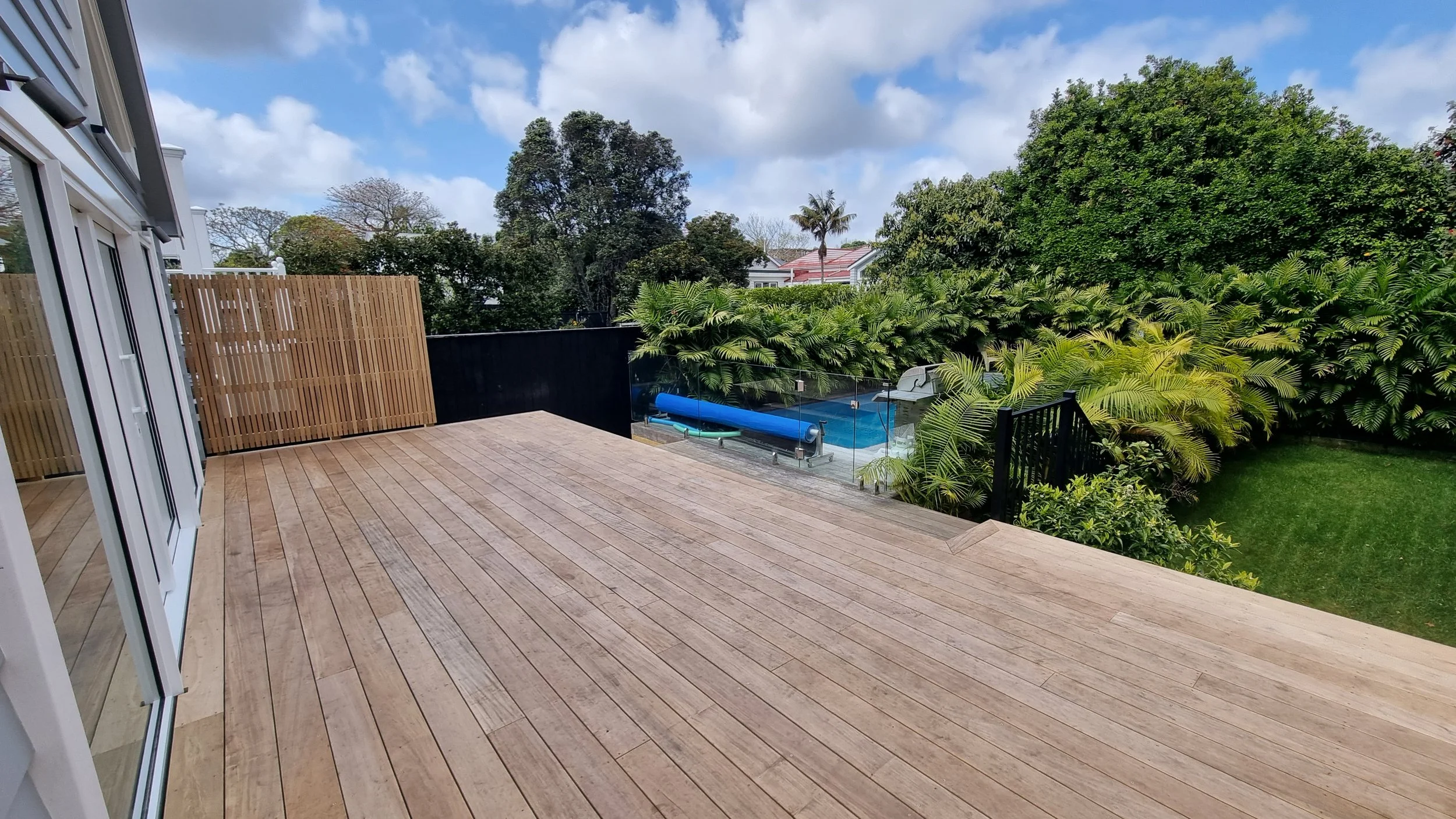 View of a backyard with a wooden deck, a swimming pool, a trampoline, lush green trees and bushes, and a partly cloudy sky.