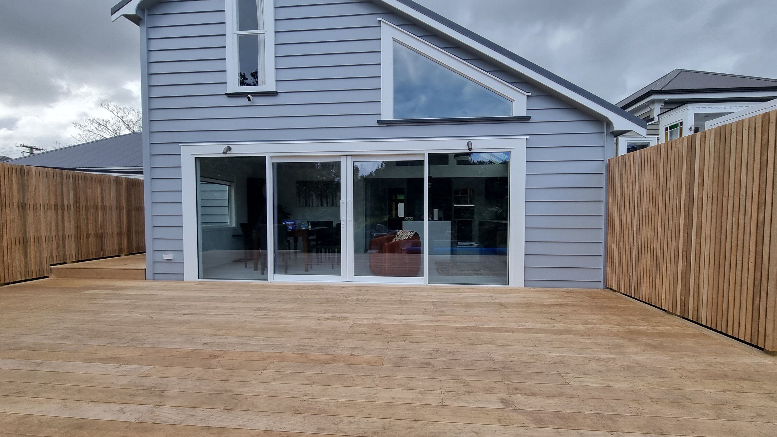 Newly finished wooden deck in backyard with large sliding glass doors leading inside a modern gray house, surrounded by wooden fencing, under cloudy sky.