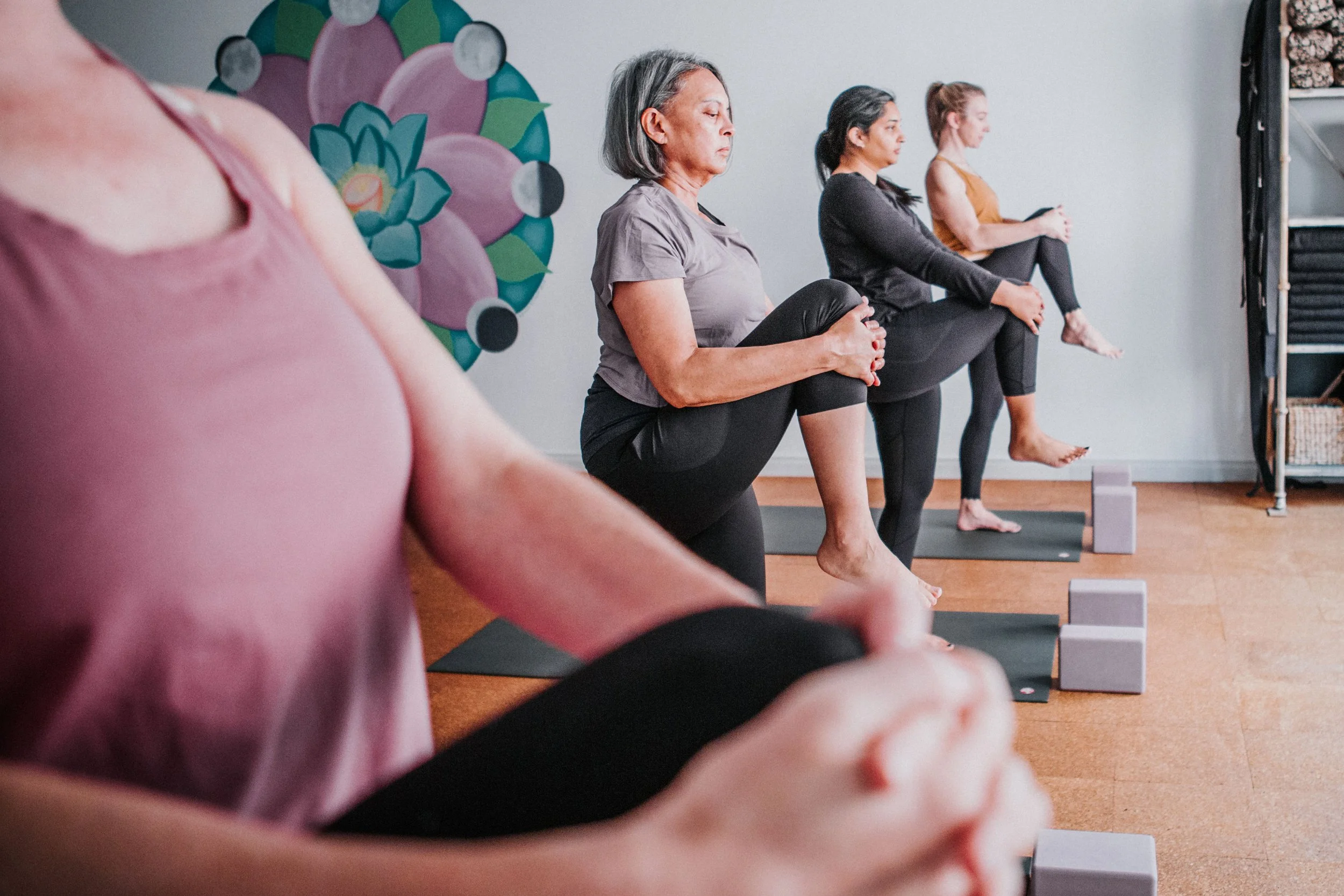 A group of women practicing yoga in a studio, sitting on yoga mats in a cross-legged pose with one leg raised, holding their knees with their hands.