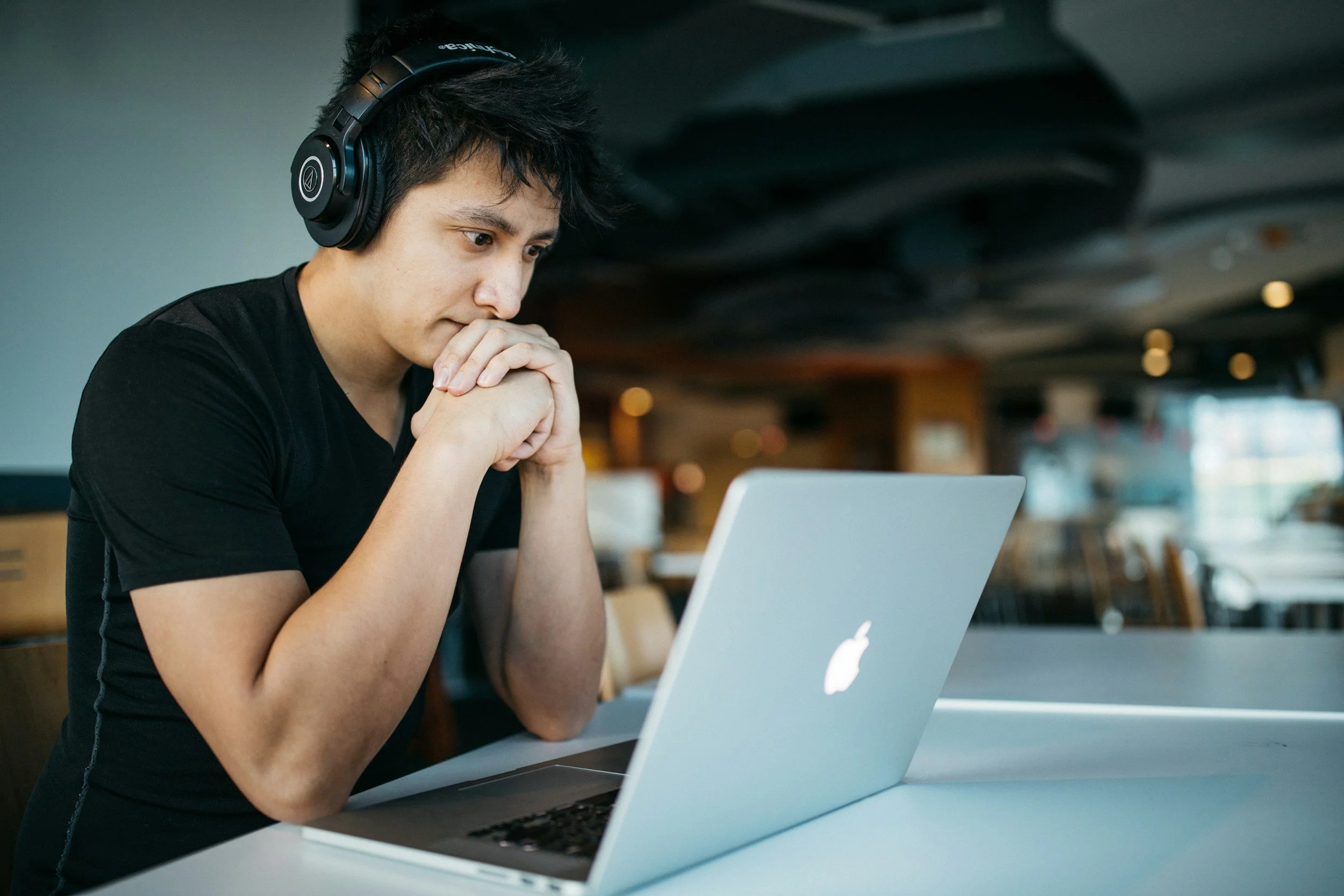 A young man with black headphones is sitting at a table, looking at a silver MacBook laptop with a focused expression in a modern, well-lit cafe.