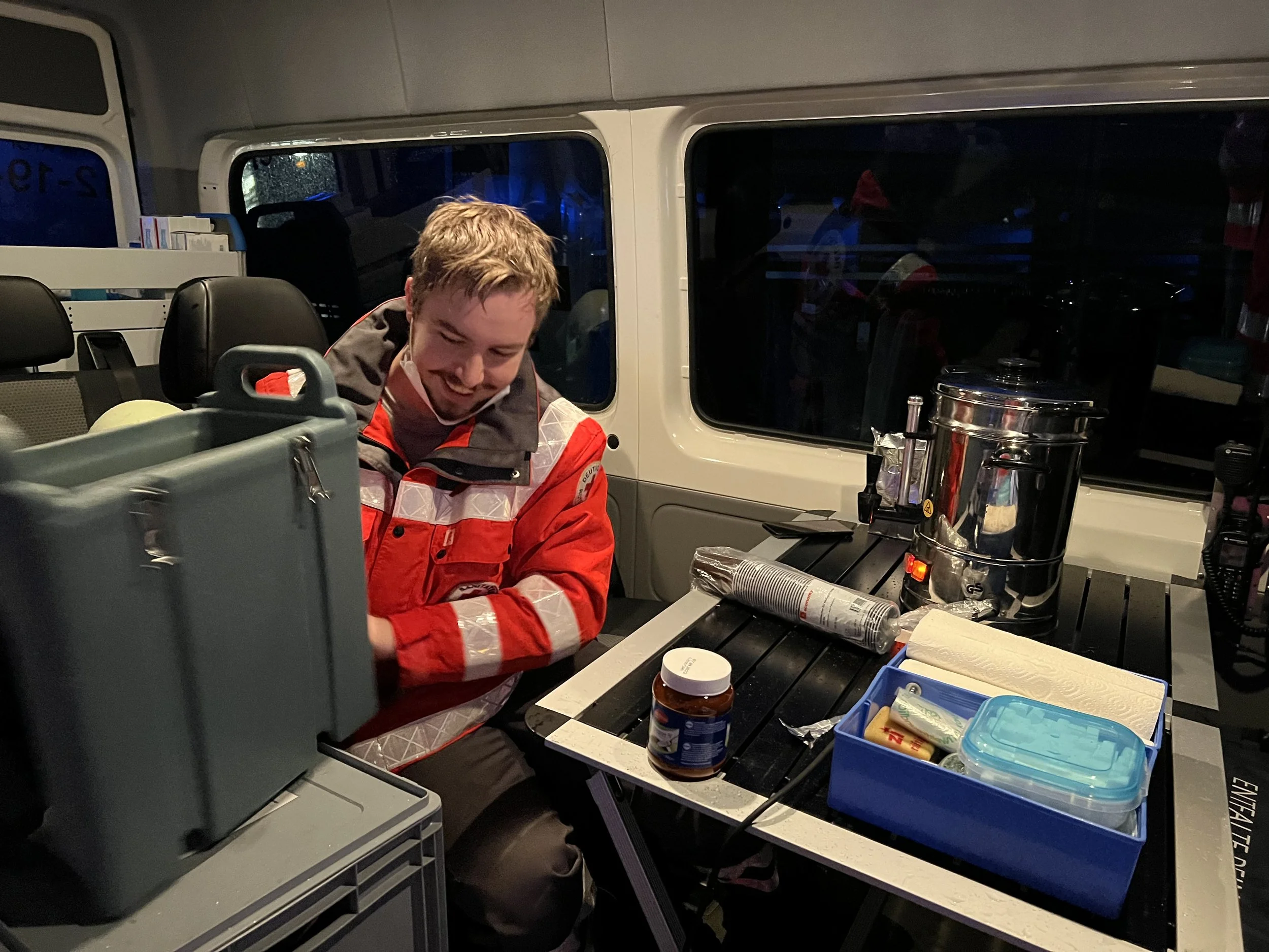 Mann in roter Jacke in einem Fahrzeug, neben einem Tisch mit Heißgetränkespender, Bechern und Lebensmittelbehältern.