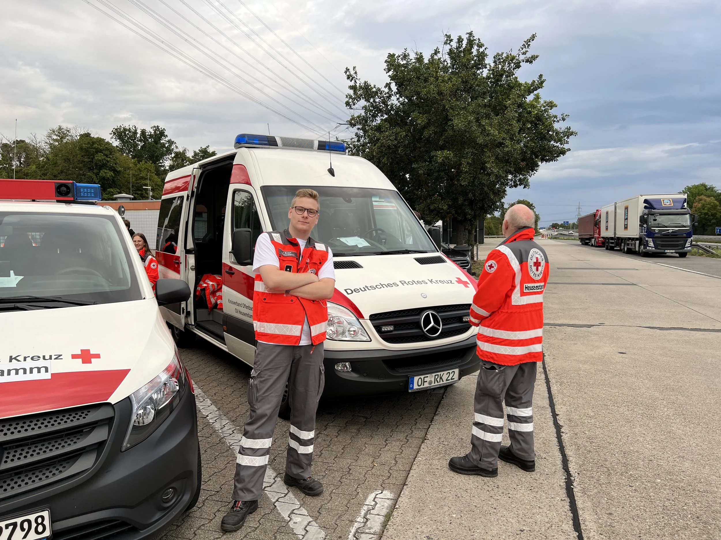 Mitglieder des Deutschen Roten Kreuzes stehen neben mehreren Rettungsfahrzeugen auf einem Parkplatz. Eine Person steht mit verschränkten Armen vor einem Fahrzeug, eine weitere Person steht daneben, während andere Mitglieder im Hintergrund sind. Mehrere Lastwagen sind in der Ferne aufgereiht.