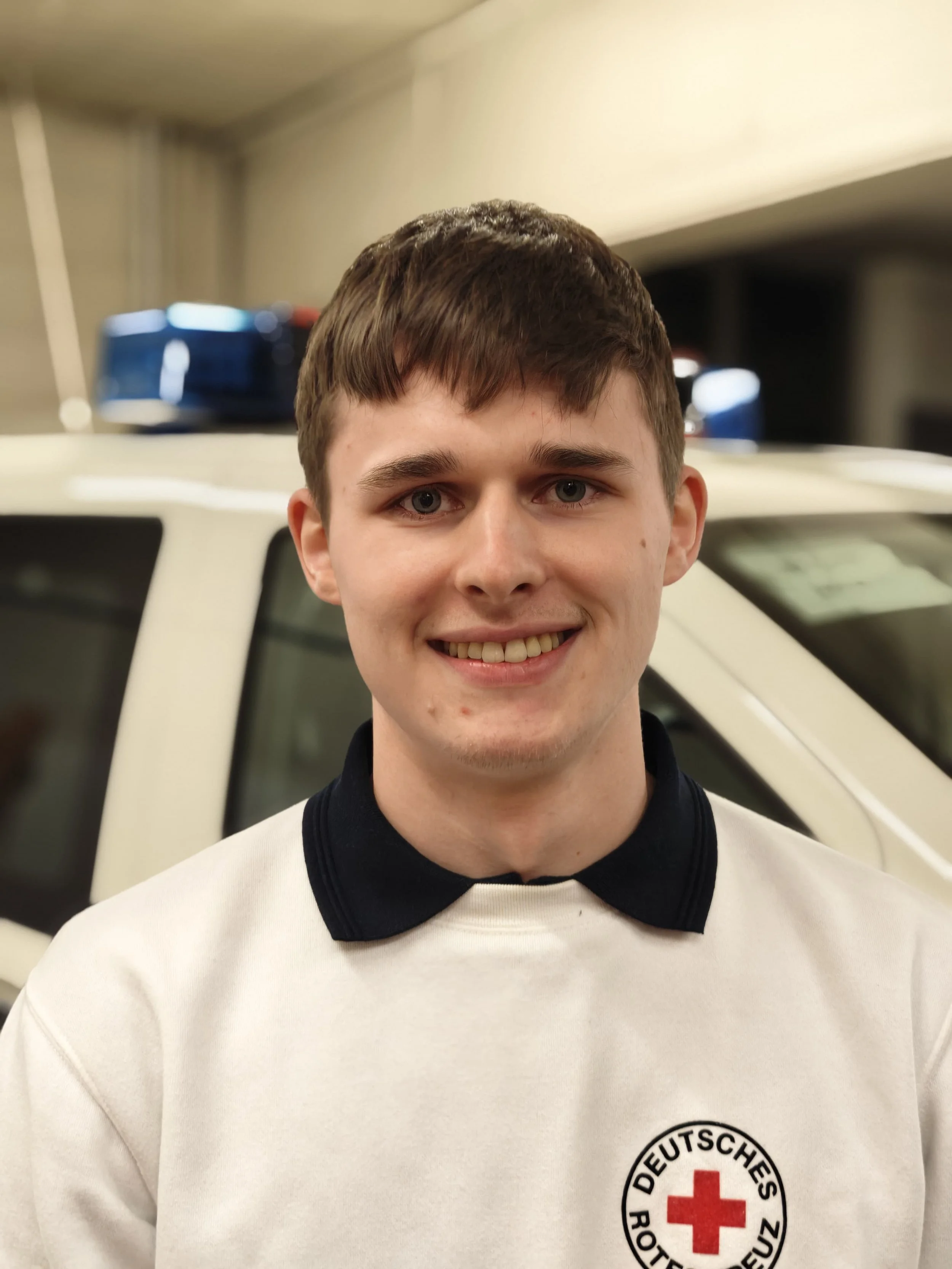 Ein junger Mann in einem Weiß-Poloshirt mit einem Das deutsche Rote Kreuz Logo, steht vor einem Rettungsfahrzeug.