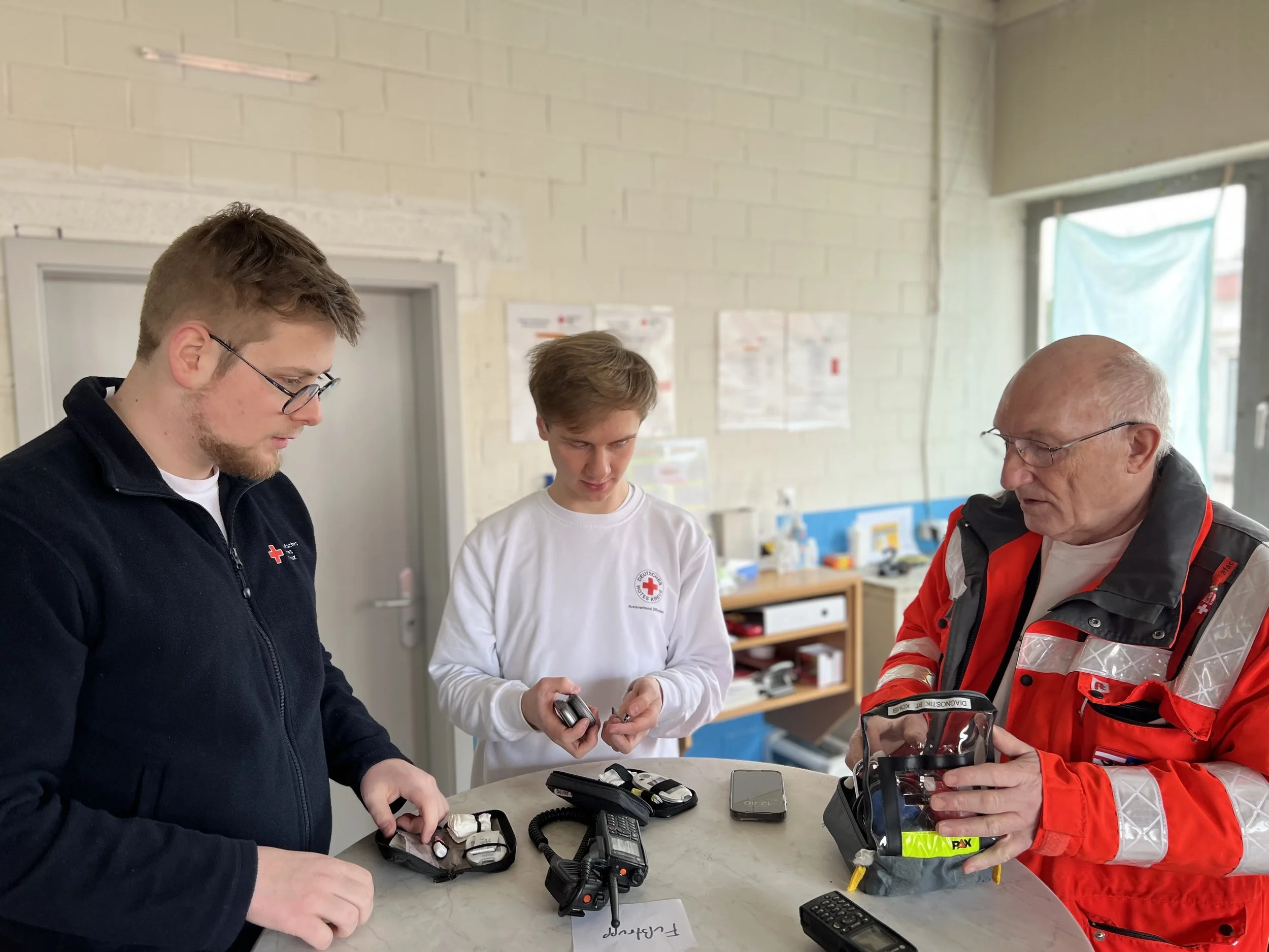 Drei Männer in Diskussion, zwei in Freizeitkleidung und einer in einer roten Rettungsdienst-Uniform. Sie stehen an einem Tisch mit Funkgeräten und anderem Equipment.