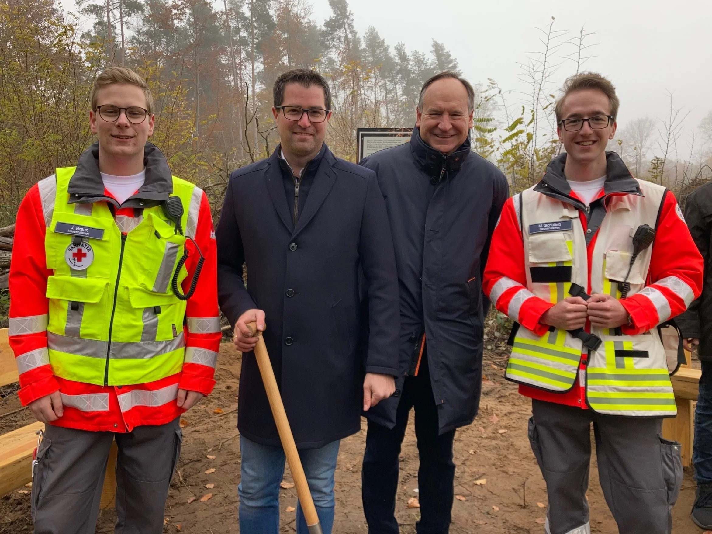 Vier Männer stehen in einer Waldumgebung. Zwei von ihnen tragen Rettungsdienstuniformen und Warnwesten des Deutschen Roten Kreuzes. Die anderen beiden tragen dunkle Mäntel. Einer der Männer hält eine Schaufel.