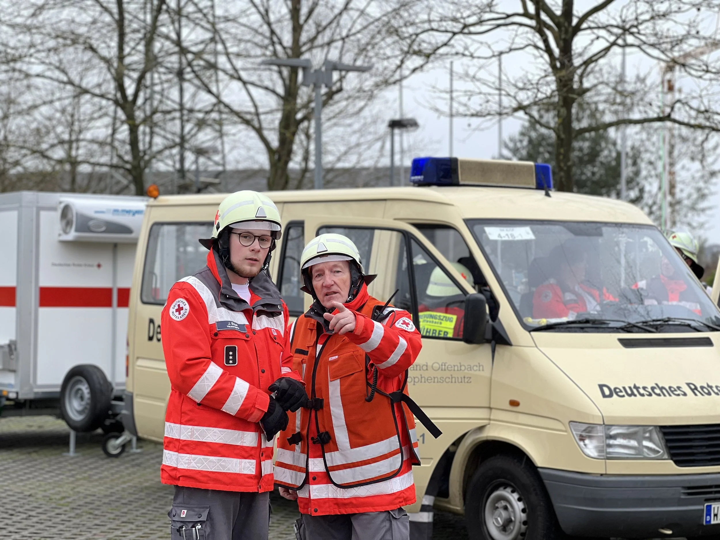 Zwei Personen in Rotkreuz-Uniformen stehen vor einem Deutschen Roten Kreuz Fahrzeug. Einer der Personen zeigt in eine Richtung. Im Hintergrund sind weitere Rettungsfahrzeuge und Bäume sichtbar.