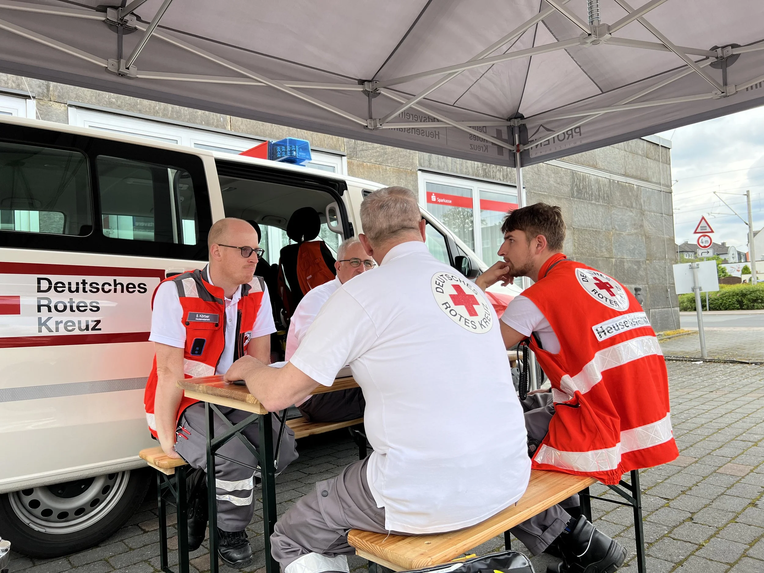 Vier Personen, die Kleidung des Deutschen Roten Kreuzes tragen, sitzen an einem Holztisch unter einem Zelt vor einem Einsatzfahrzeug.